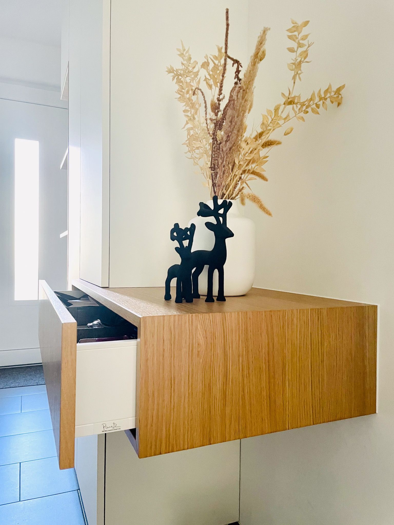Schwebendes Holzregal mit offener Schublade; schwarze Hirschfiguren und Trockenblumen in einer weißen Vase.