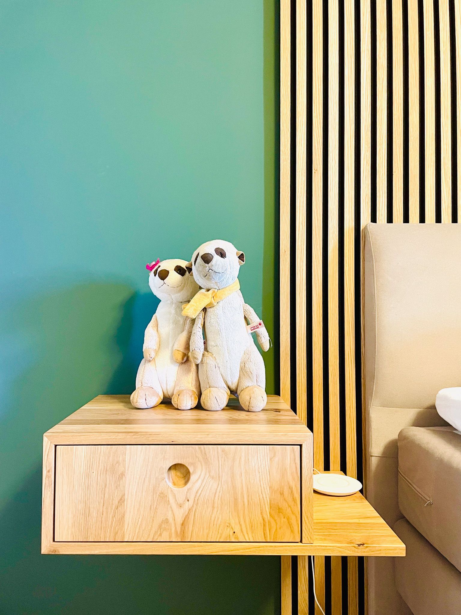 Zwei Erdmännchen-Stofftiere auf einem hölzernen Nachttisch neben einem Bett mit grüner Wand und Holzvertäfelung.