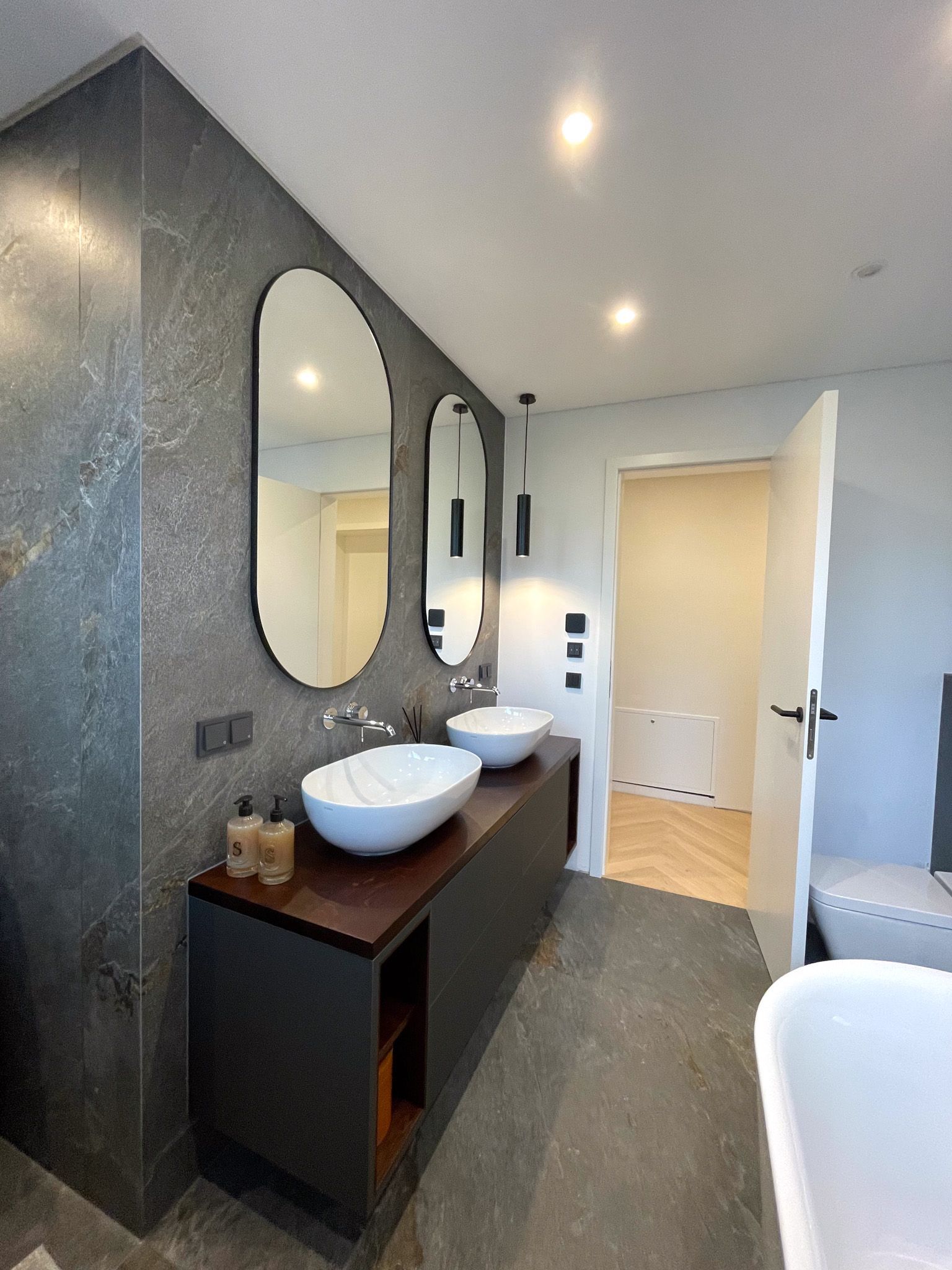 Modernes Badezimmer mit dunkelgrauen Wänden, zwei ovalen Spiegeln und weißen Waschbecken auf einem schwarzen Waschtisch.