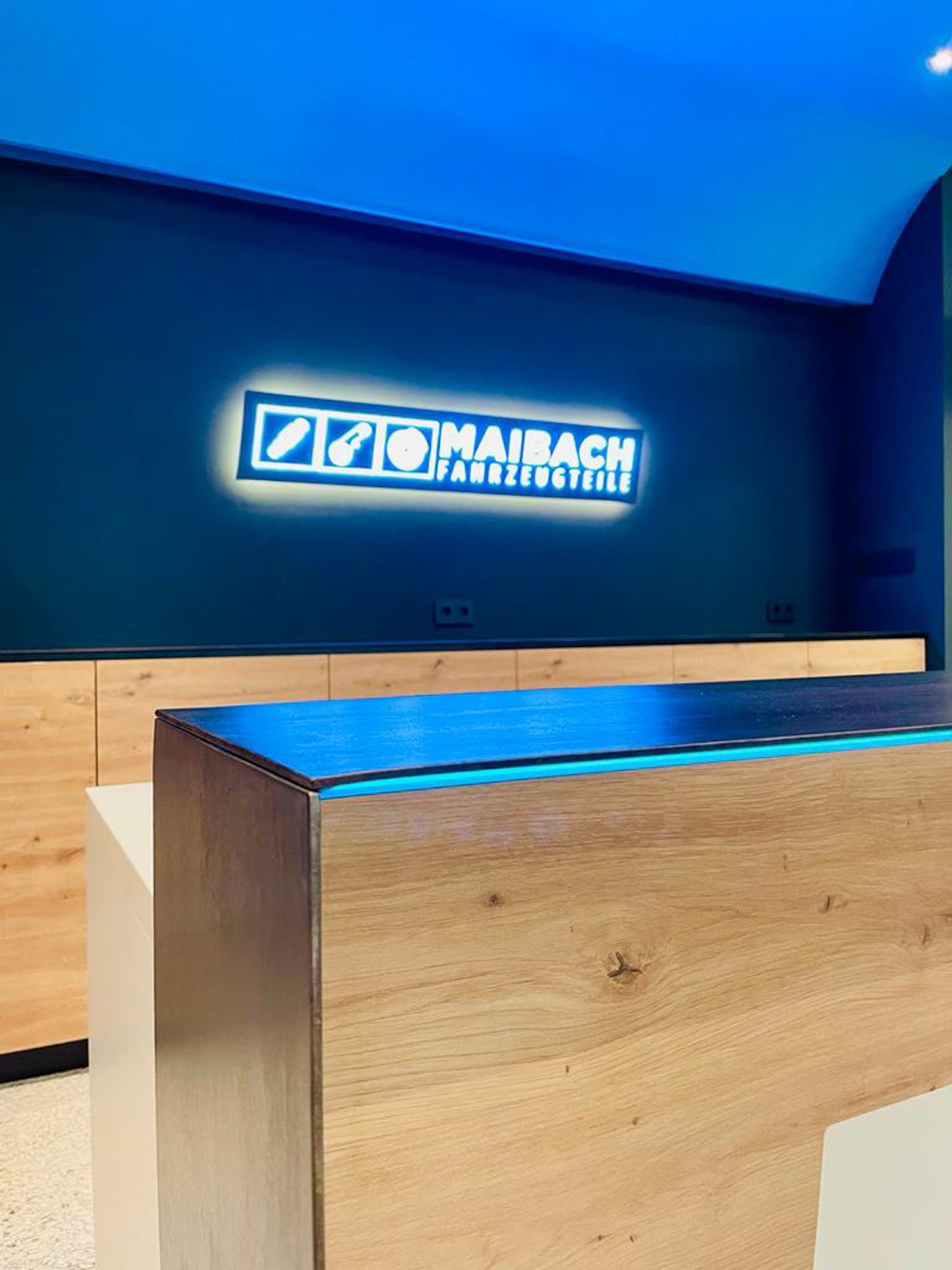 Holztheke mit beleuchtetem Logo „MAIBACH FARBPOTRIE“ auf blauer Wand.