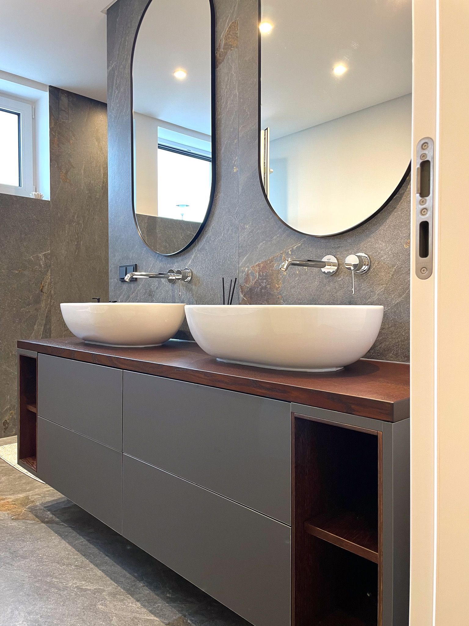 Badezimmer mit grauem Waschtisch, zwei ovalen Spiegeln und weißen Waschbecken.