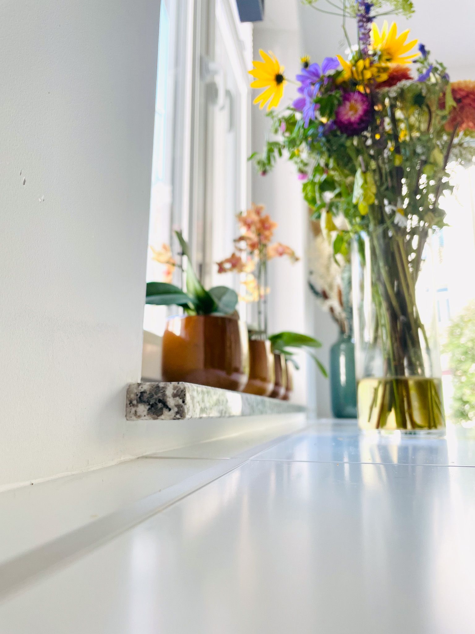 Weiße Fensterbank mit Topfpflanzen und einer Vase mit bunten Blumen.