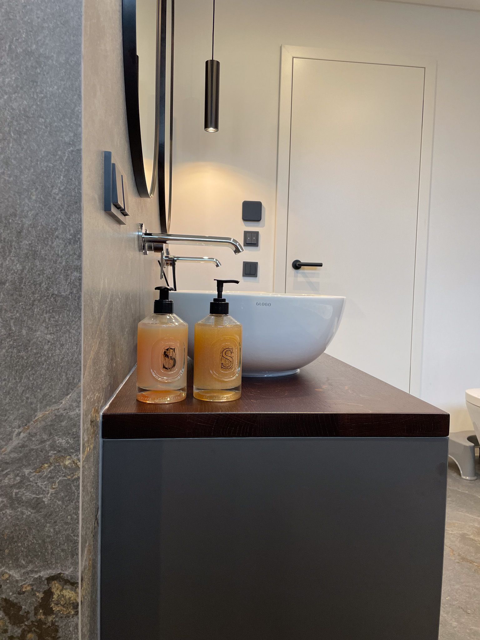 Badezimmerwaschtisch mit rundem Waschbecken, Seifenspendern und einer dunklen Arbeitsplatte vor einem grauen Schrank.