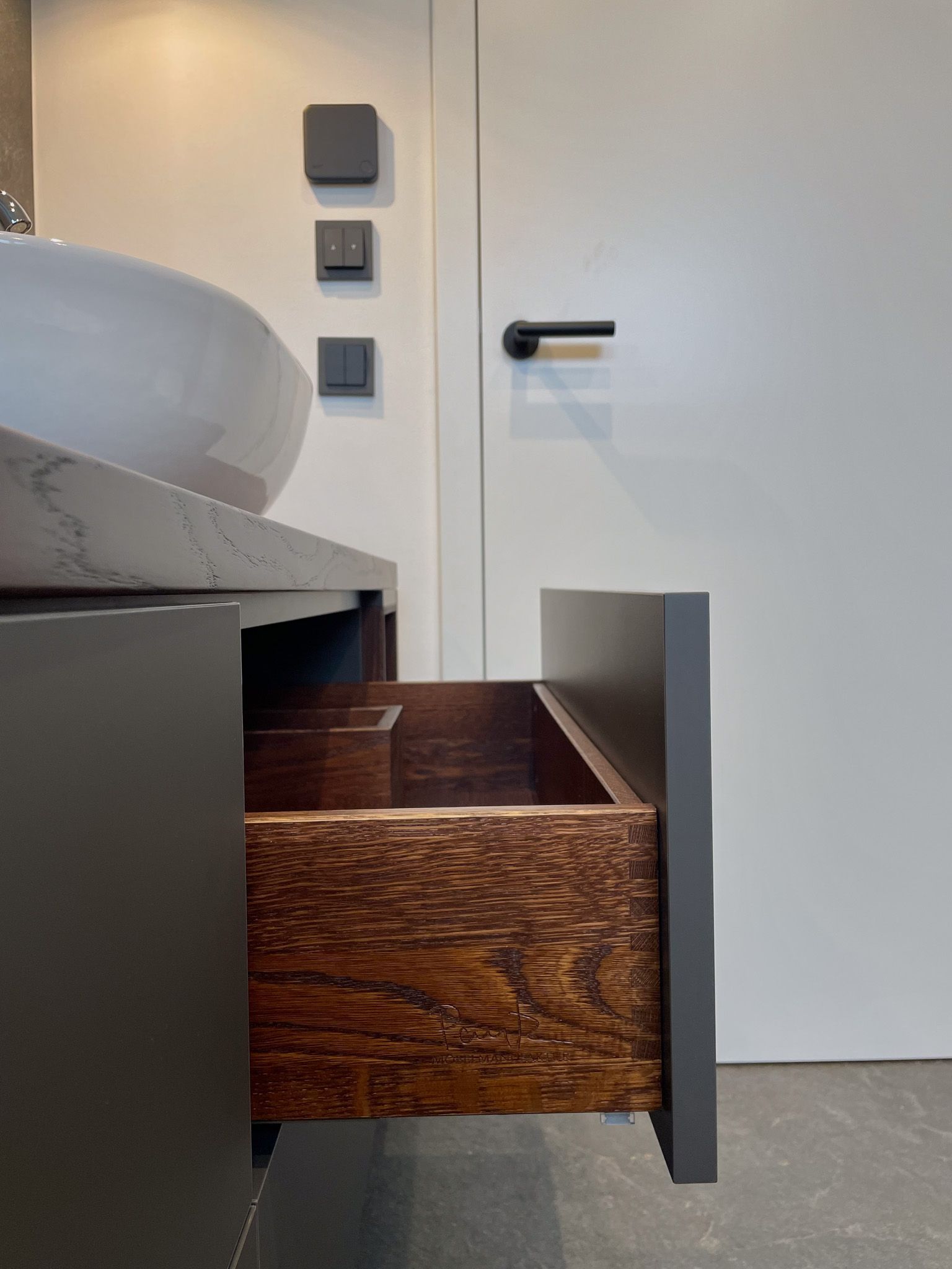 Badezimmerwaschtisch mit offener Holzschublade, grauer Arbeitsplatte, weißem Waschbecken und Tür.