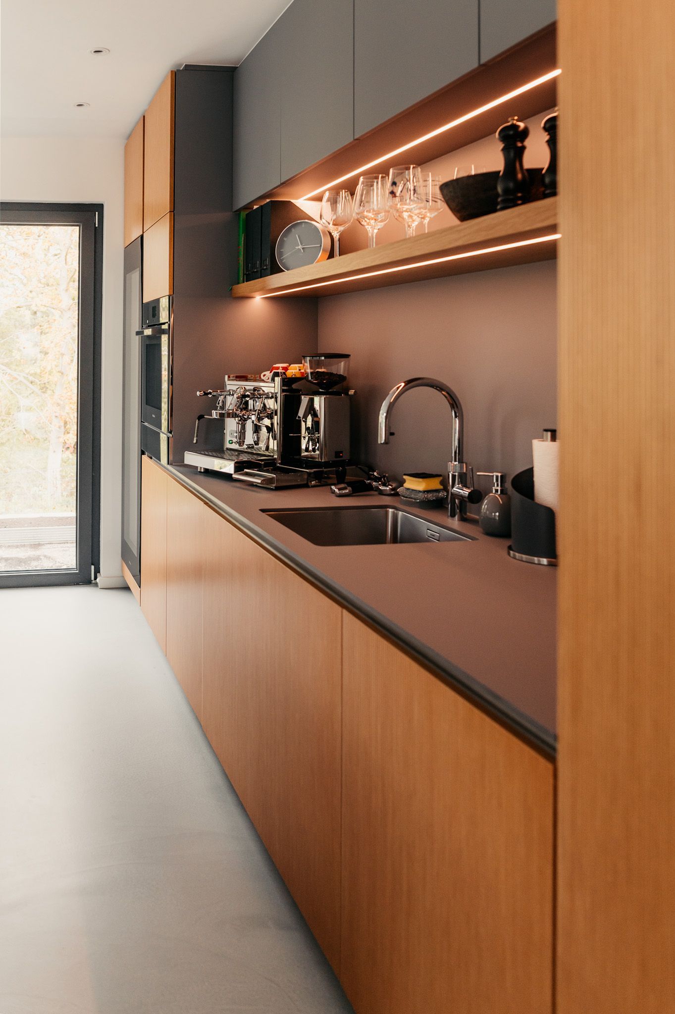 Moderne Küche mit Holzschränken, grauer Küchenrückwand und Deckenbeleuchtung; die Geräte stehen auf der Arbeitsplatte.