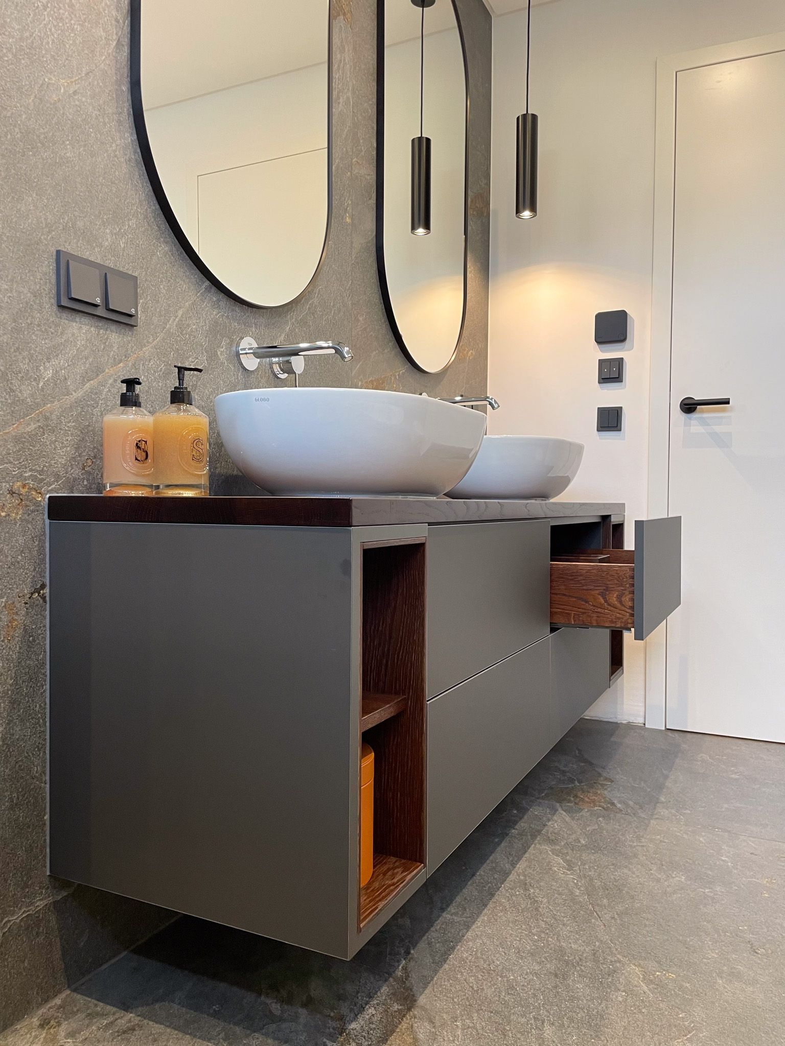 Modernes Badezimmer mit grauem schwebendem Waschtisch, weißen Aufsatzwaschbecken, ovalen Spiegeln und braunen Akzenten.