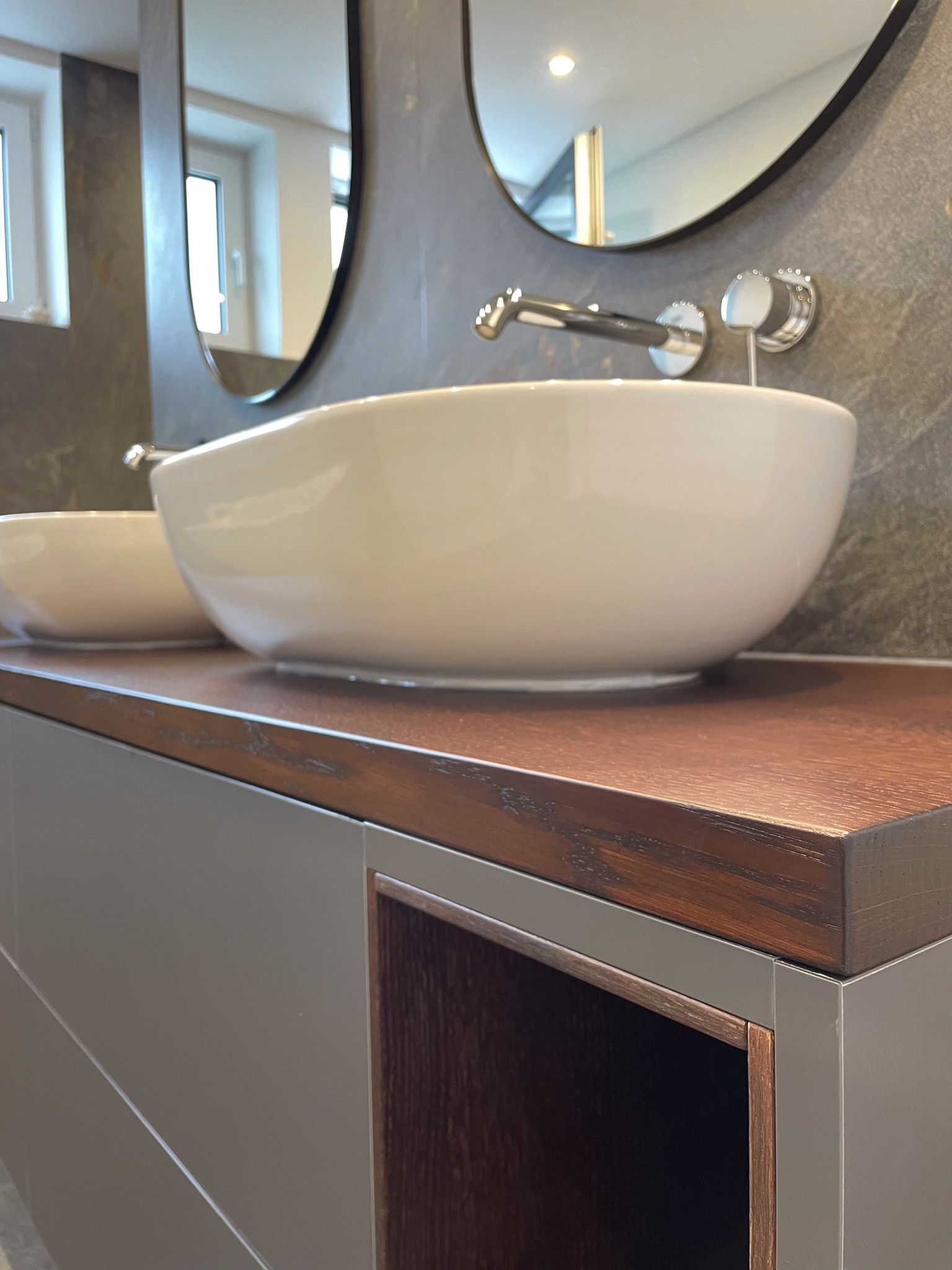 Badezimmerwaschtisch mit weißem ovalem Waschbecken, brauner Arbeitsplatte und grauen Schränken.