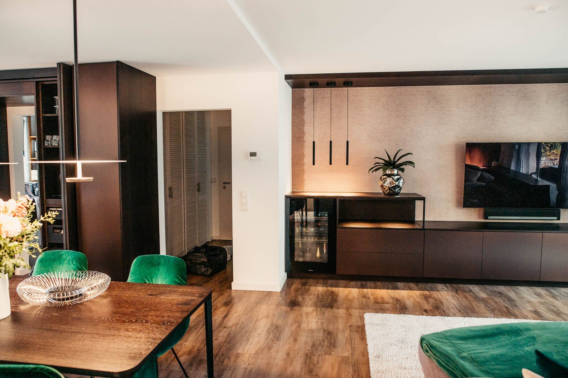 Modernes Wohnzimmer mit dunklen Holzmöbeln, grünen Samtstühlen und einem an der Wand montierten Fernseher.