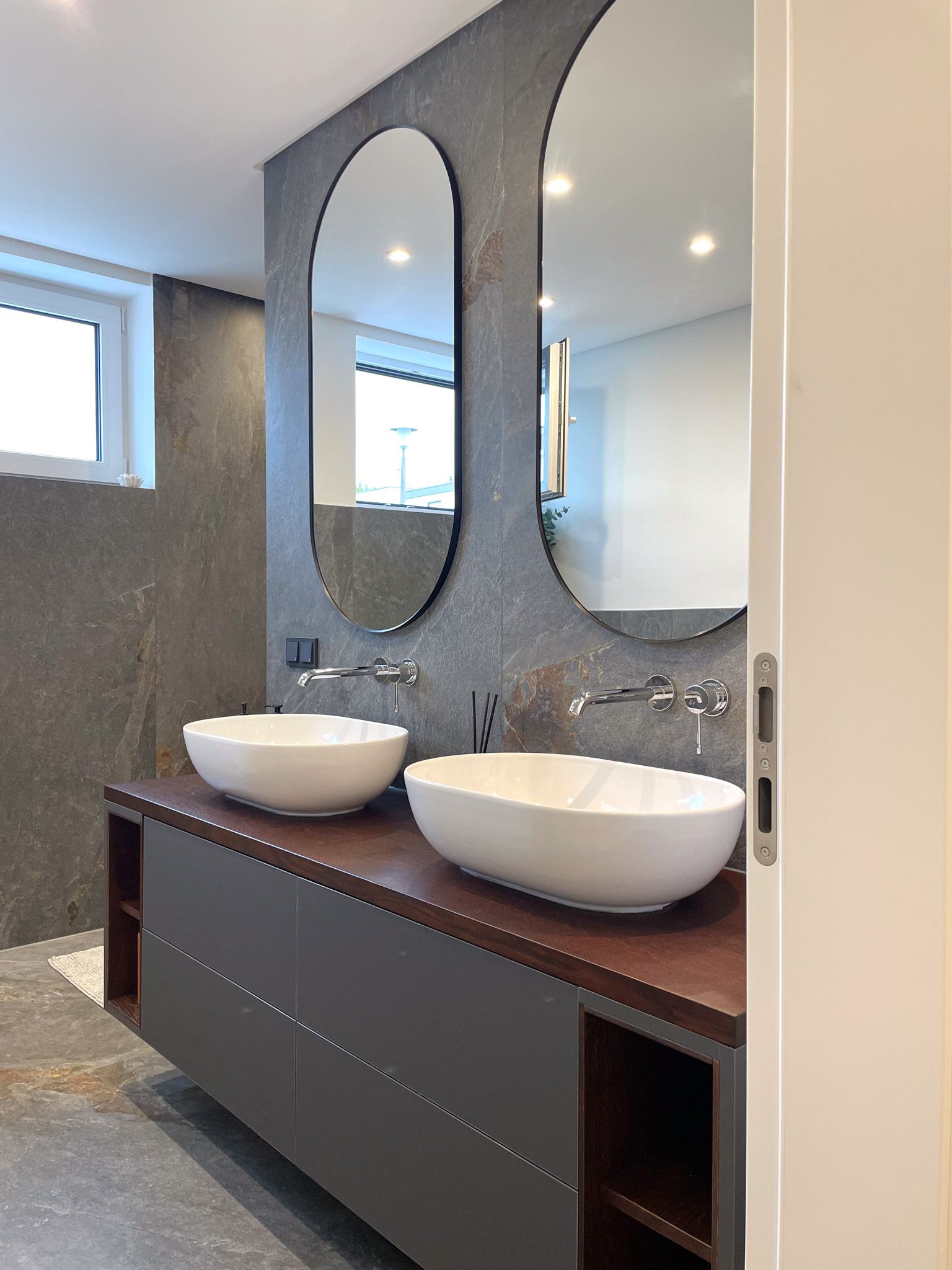 Modernes Badezimmer mit grauem Waschtisch, weißen Waschbecken, ovalen Spiegeln und grauer Steinwand.