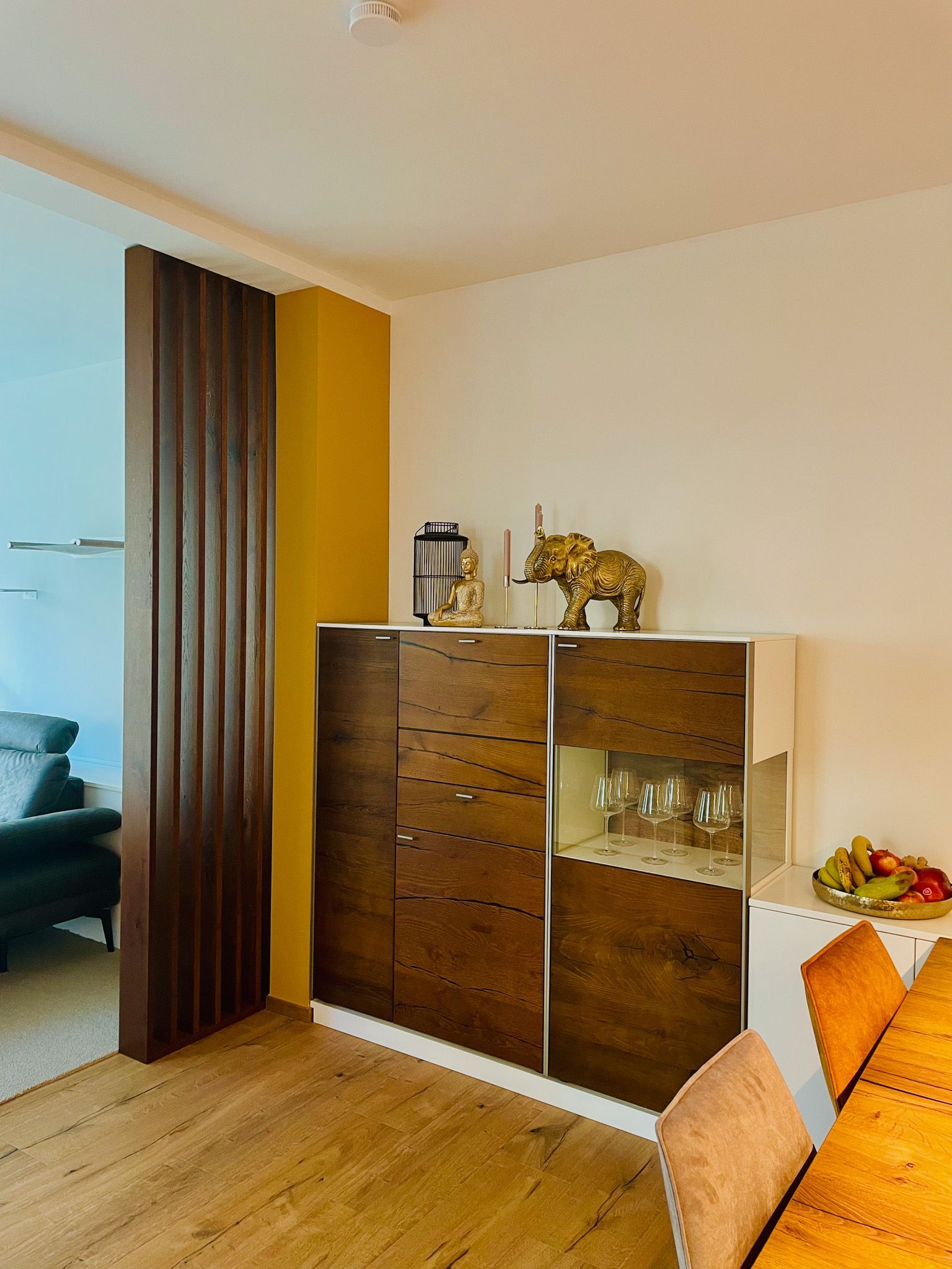 Dekorierter Holzschrank neben einer gelben Wand und einer strukturierten Trennwand in der Nähe eines Essbereichs.