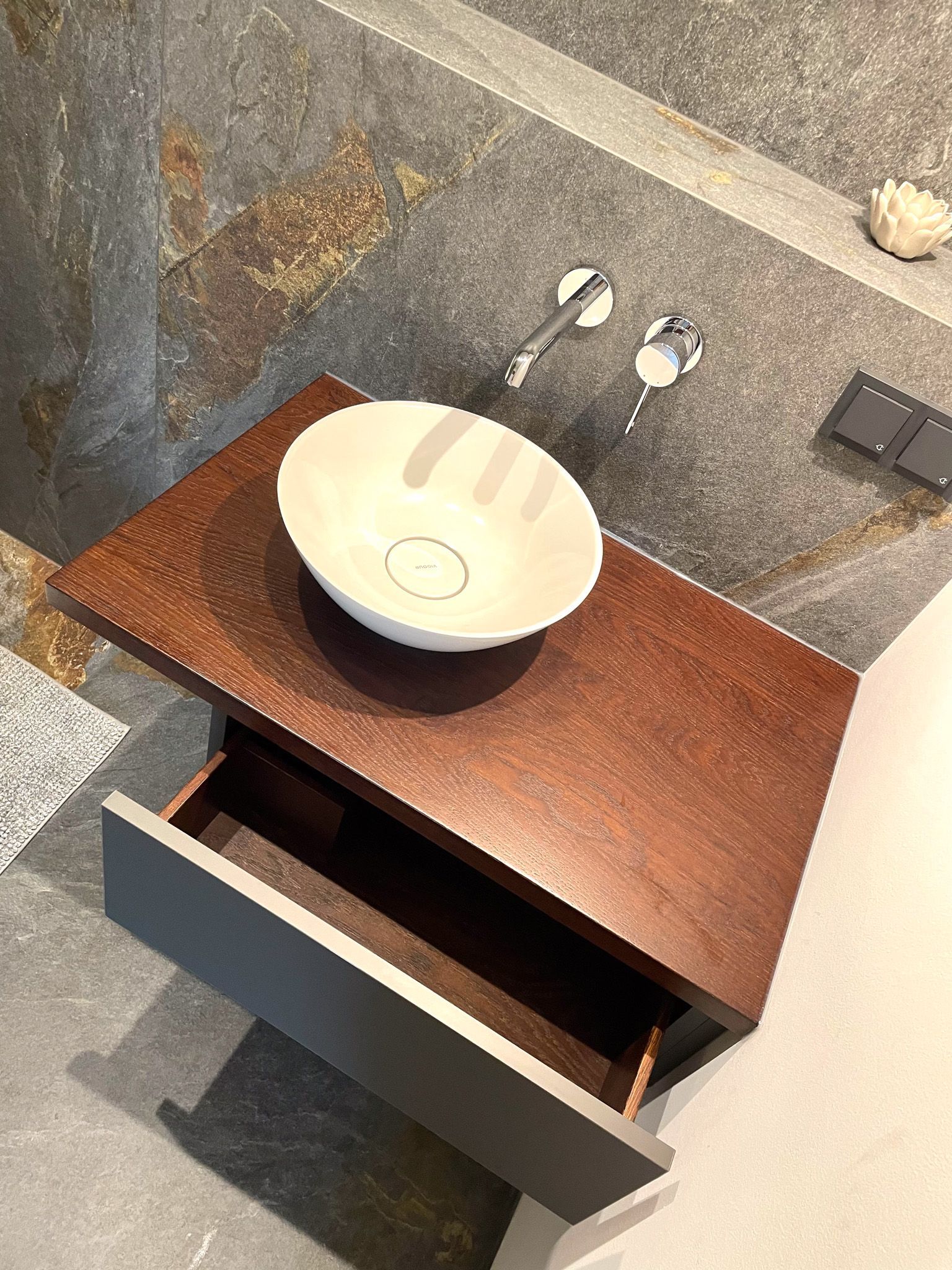 Ein moderner Badezimmerwaschtisch mit Waschbecken. Braune Arbeitsplatte, graue Schublade, Steinwand und ein weißes Aufsatzwaschbecken.