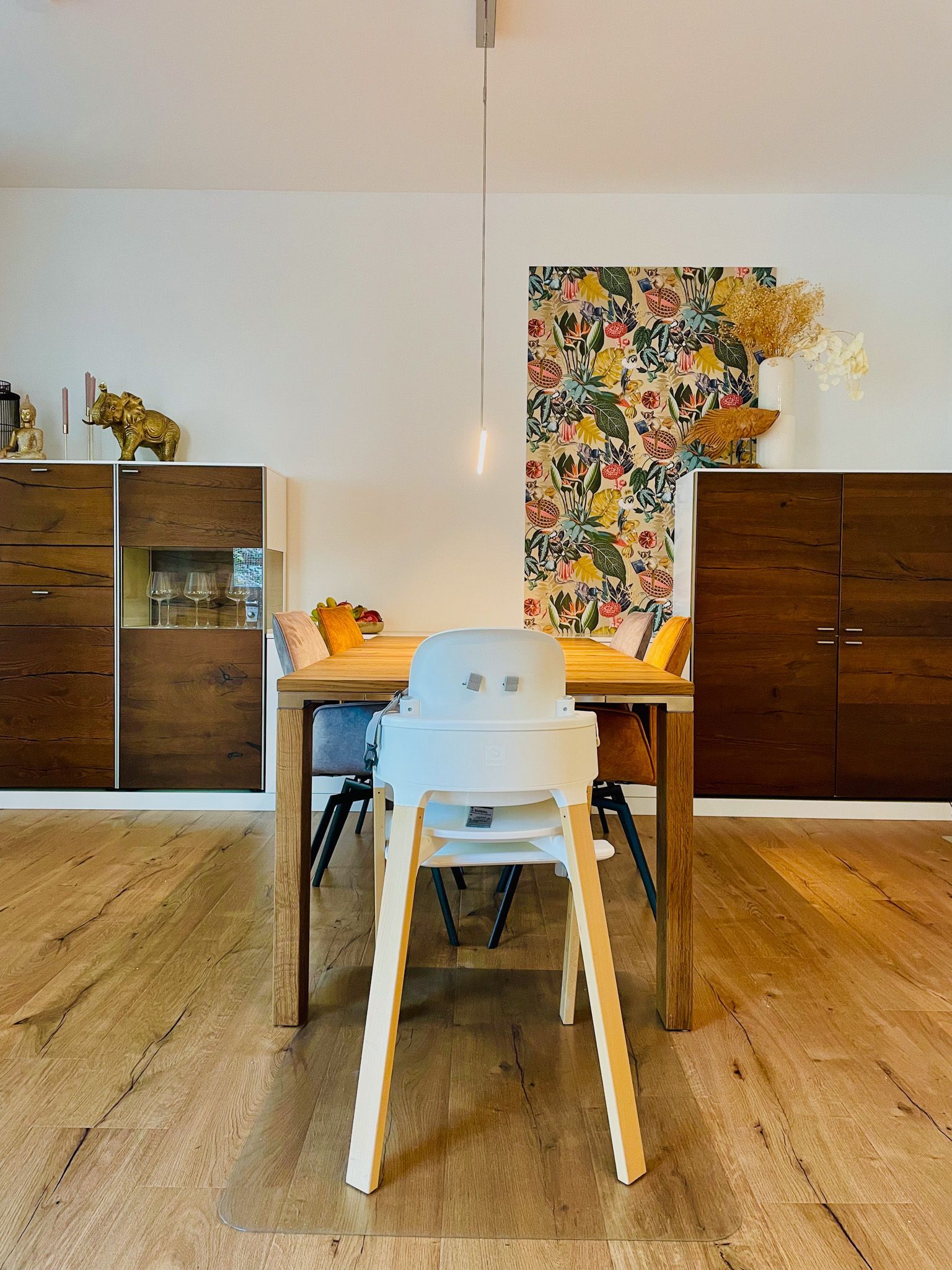 Ein heller Hochstuhl an einem hölzernen Esstisch in einem Raum mit Holzschränken und einem Blumengemälde.