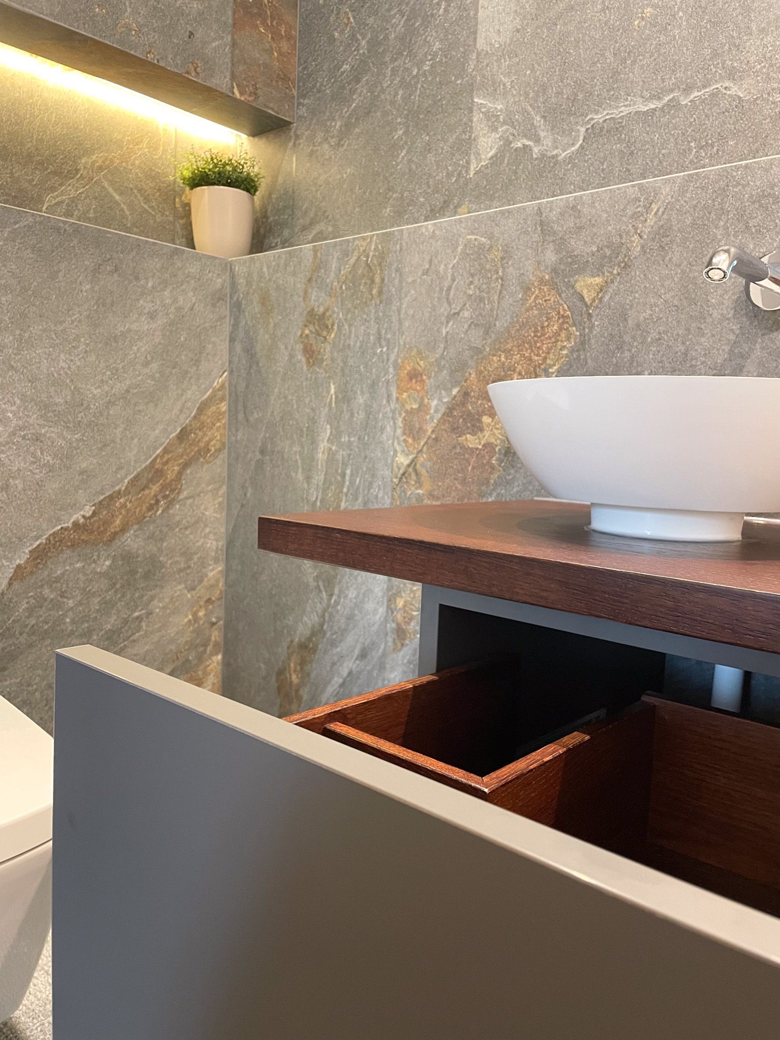 Modernes Badezimmer: Grauer Waschtisch mit offener Holzschublade, Steinwand, weißem Waschbecken und einer kleinen Pflanze.