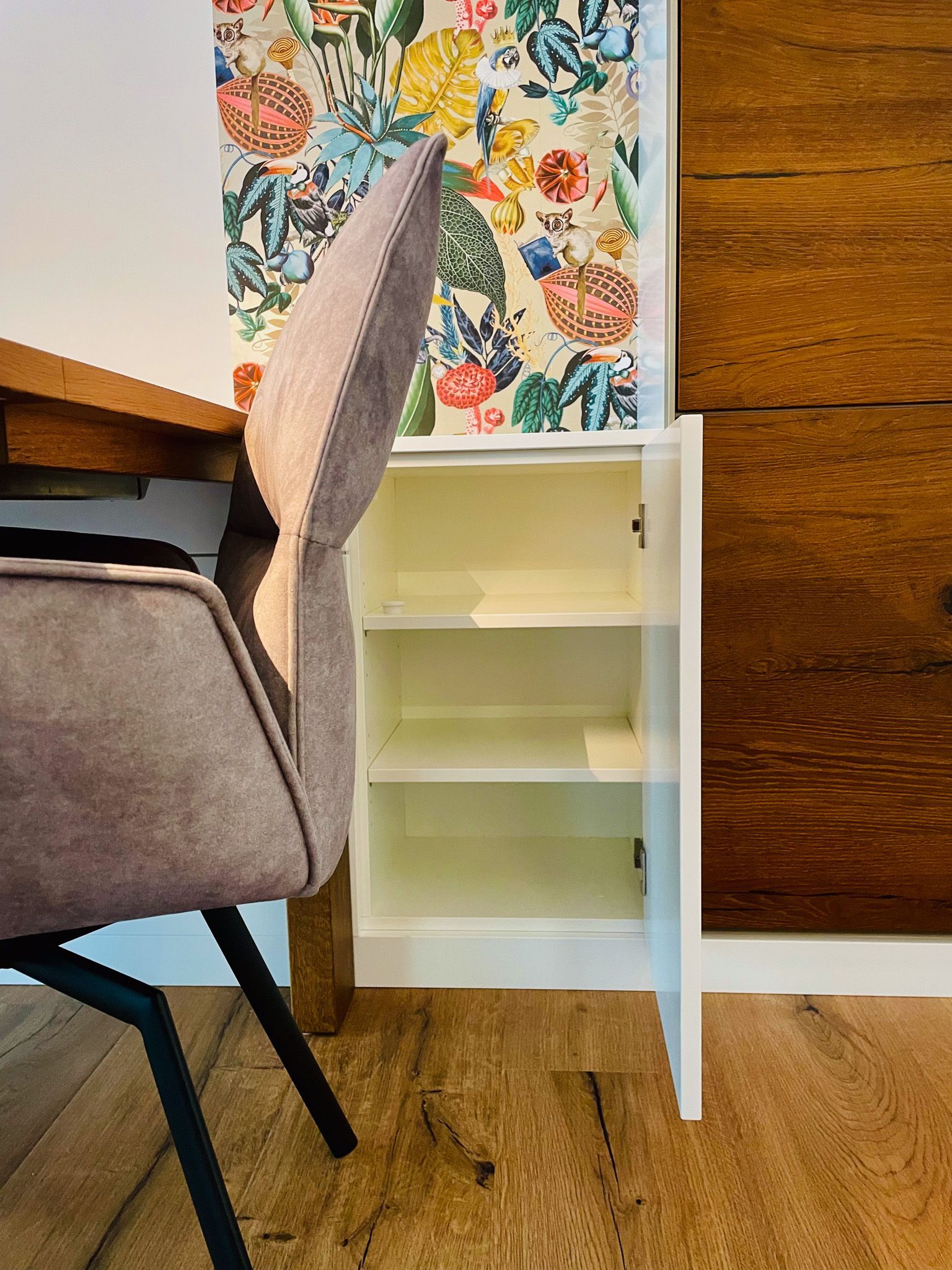 Offener weißer Schrank mit Regalen, daneben eine gemusterte Tapete und ein Stuhl an einem Holztisch.