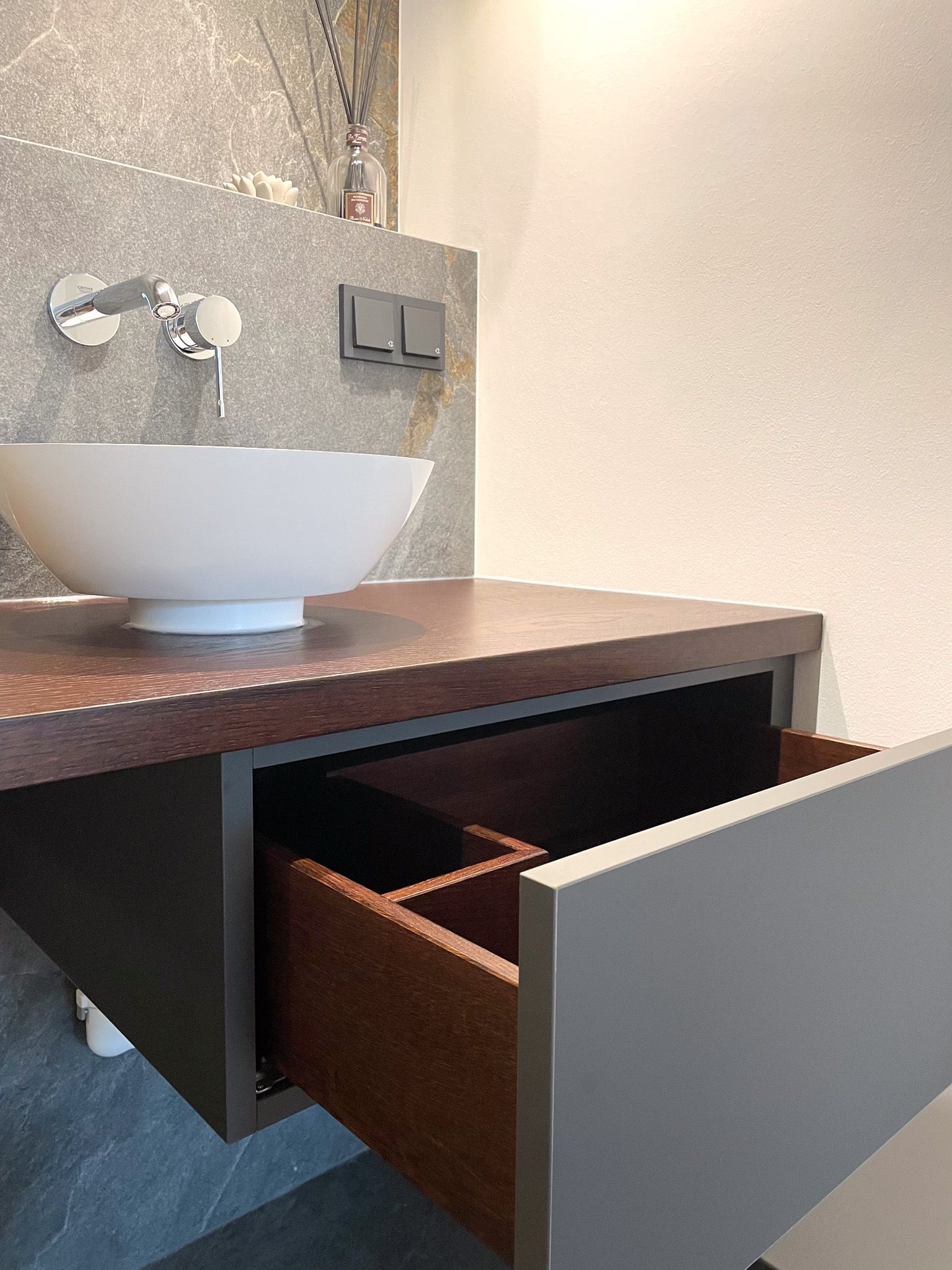 Schwebender Badezimmerwaschtisch mit offener Schublade; weißes Waschbecken, braune Arbeitsplatte und Schubladeninnenseiten, graue Schubladenfronten.
