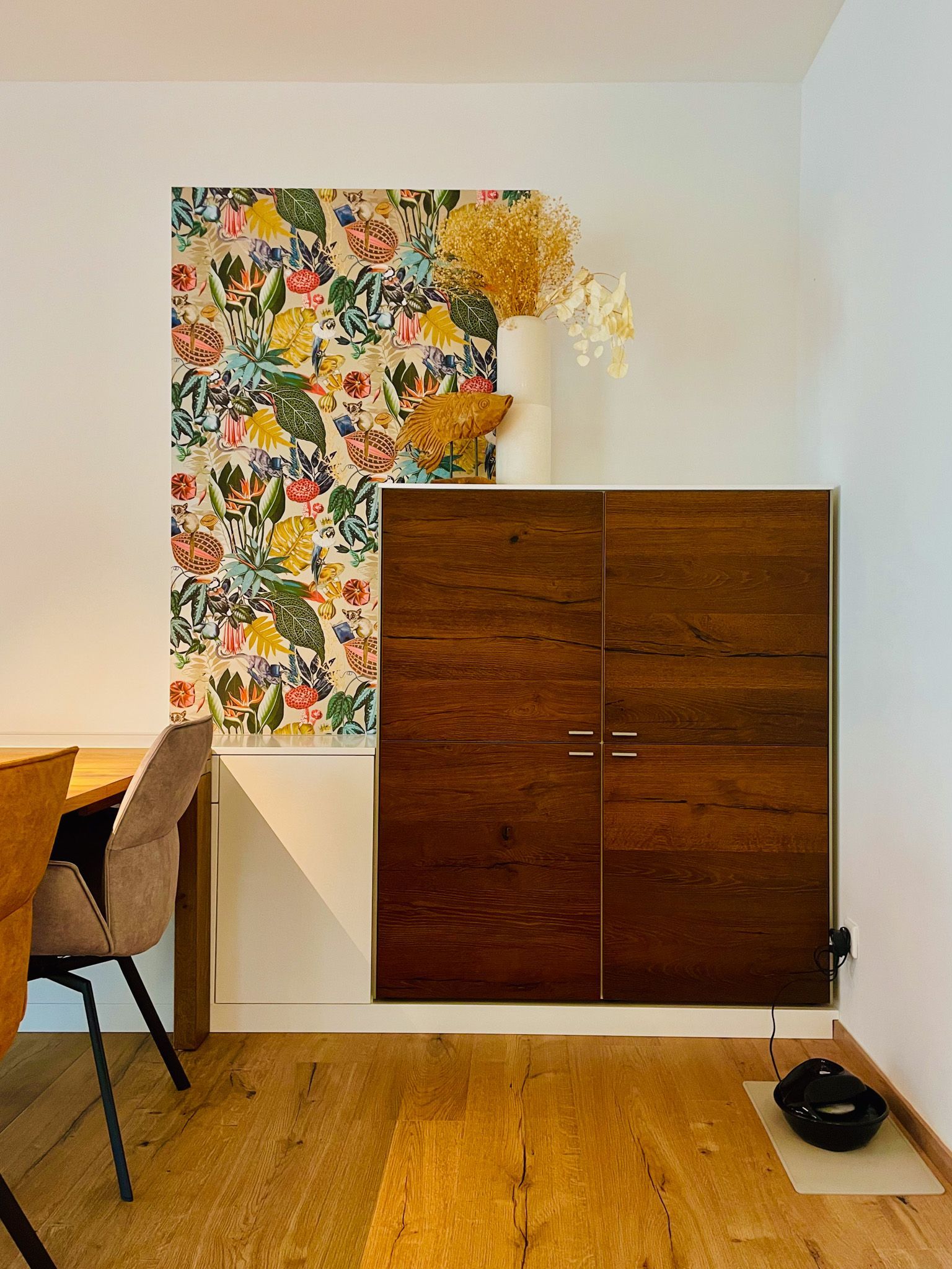 Schrank mit dunklen Holztüren, geblümter Tapete und Vase mit Trockenblumen.