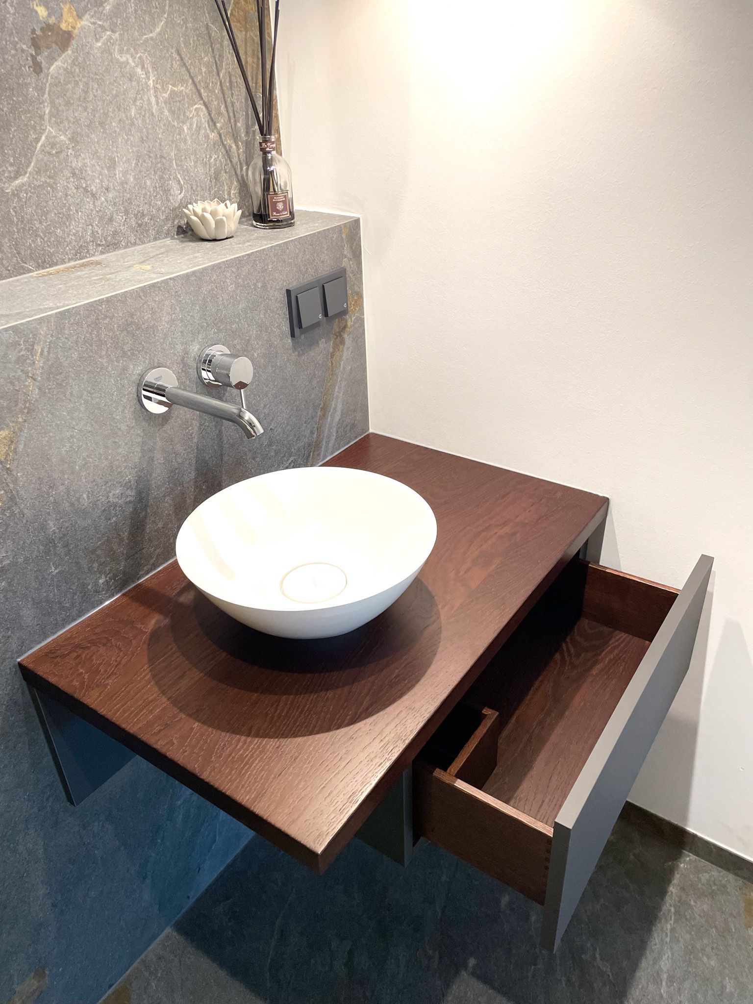 Ein schwebender Badezimmerwaschtisch mit einem runden weißen Waschbecken, einer braunen Arbeitsplatte und einer offenen Schublade; vor einer grauen Steinwand.