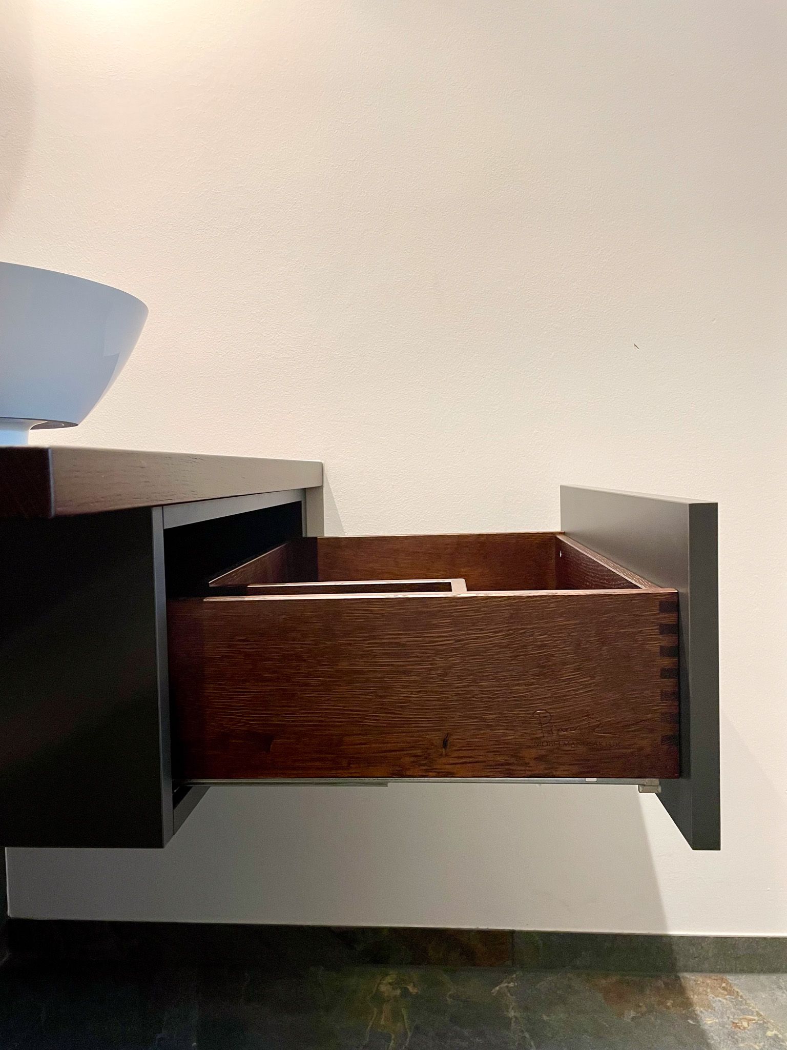 Aus einem schwebenden Badezimmerwaschtisch vor einer weißen Wand wird eine dunkel gebeizte Holzschublade herausgezogen.