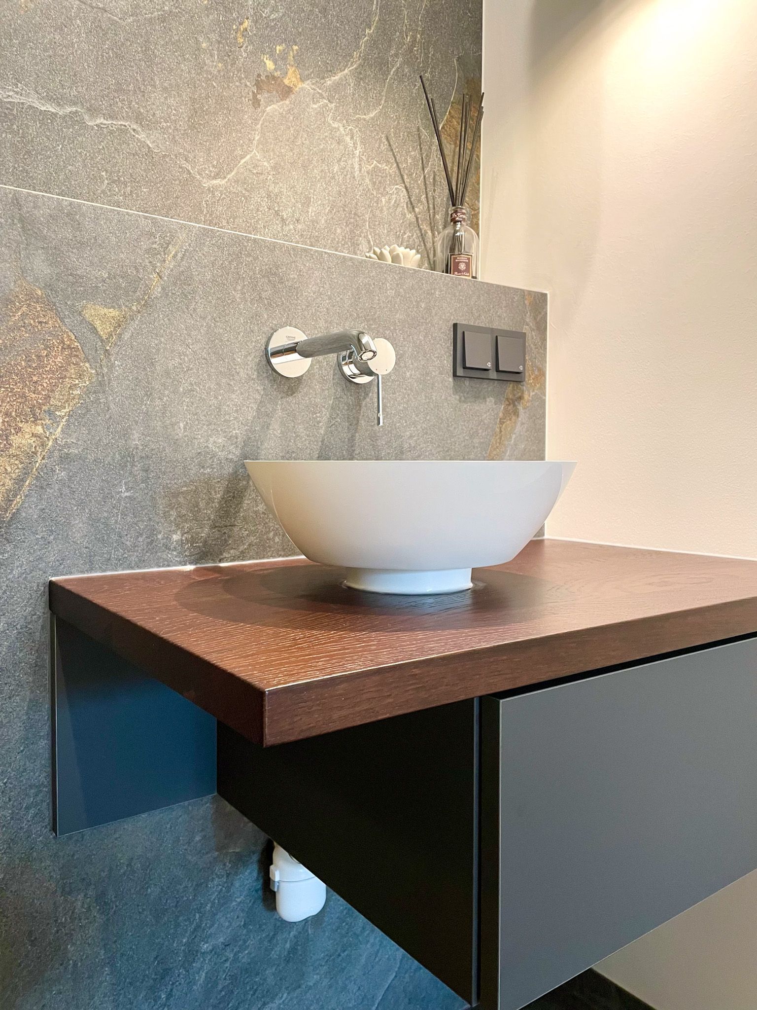 Modernes Badezimmer mit Aufsatzwaschbecken auf einer braunen Ablage vor einer strukturierten grauen Wand. Darunter ein dunkelgrauer Schrank.