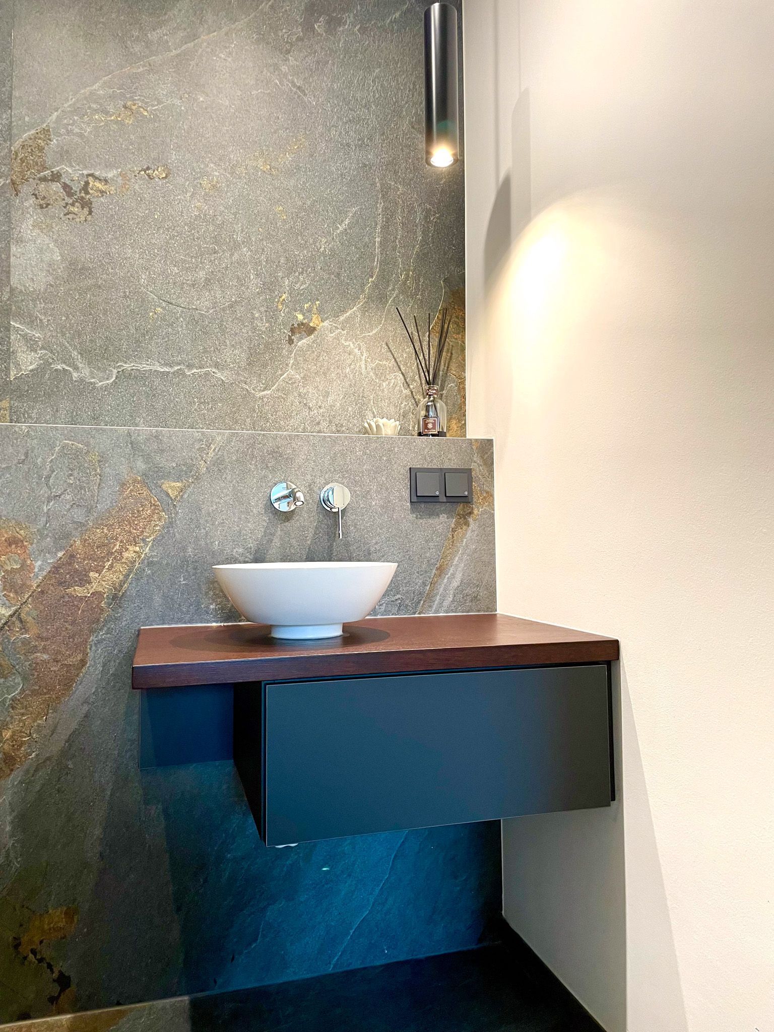 Schwebender Badezimmerwaschtisch mit weißem Waschbecken, dunklem Schrank und strukturierter Steinwand.