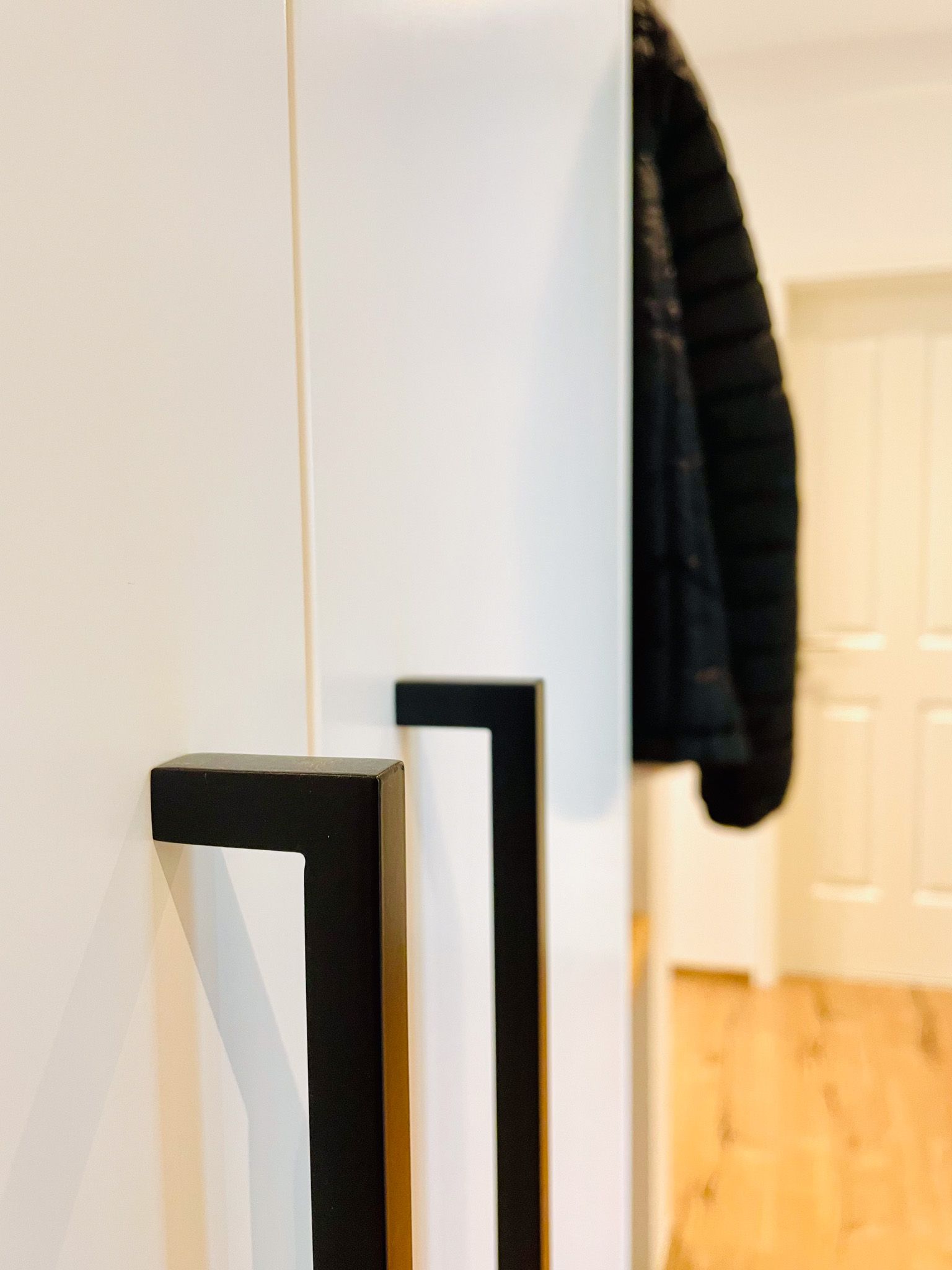 Weißer Schrank mit schwarzen Griffen, seitlich hängt eine dunkle Jacke.