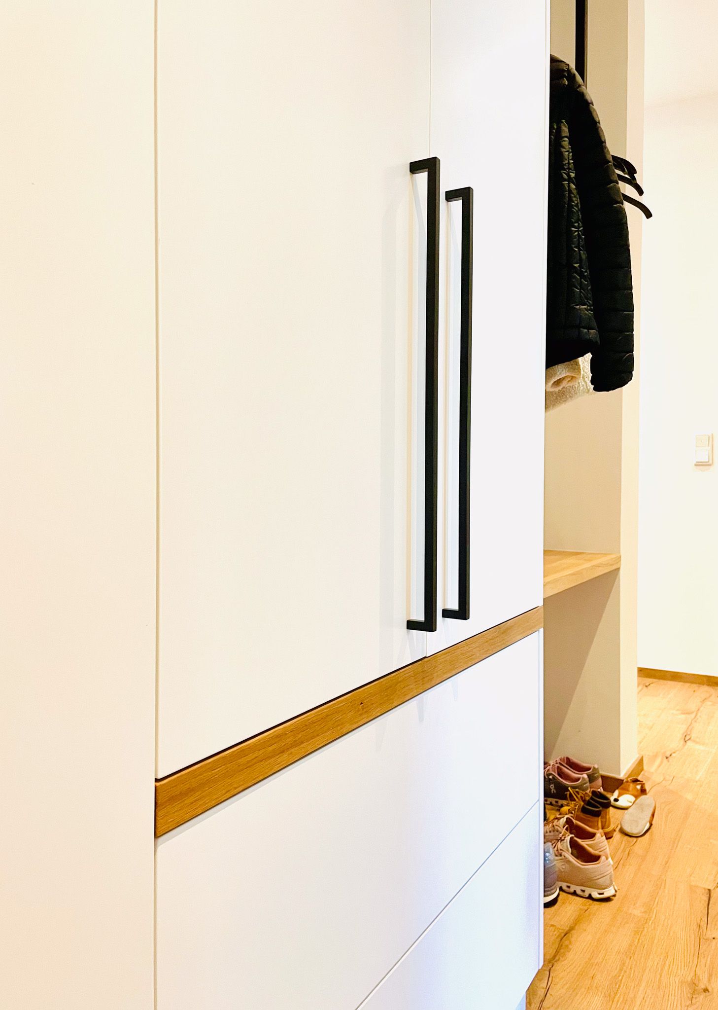 Weißer Schrank mit schwarzen Griffen und Holzregal, Mantel und Schuhe sichtbar.