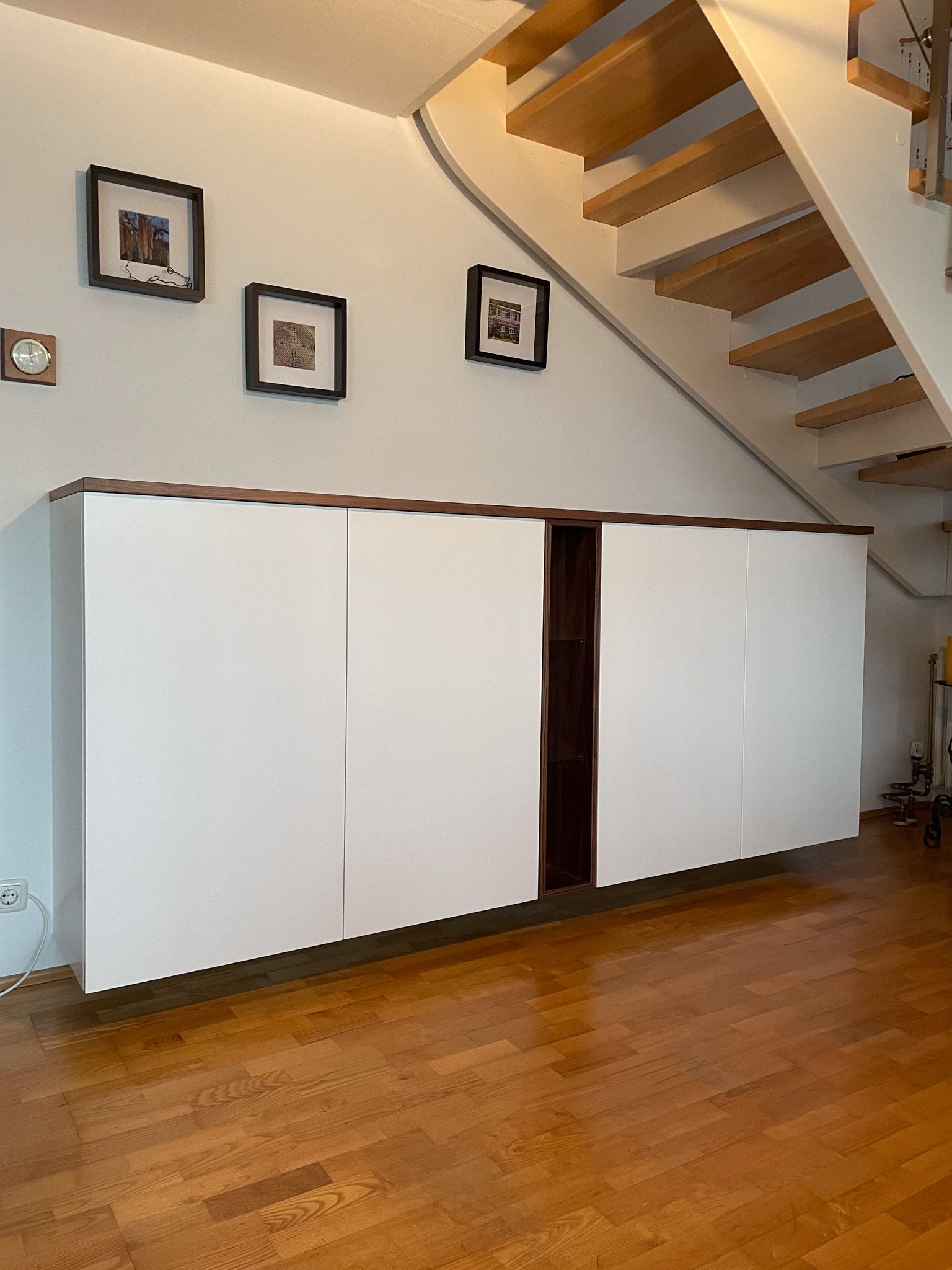 Weißer Schrank mit braunen Zierleisten unter einer Treppe. Bilder an der Wand, Holzboden.