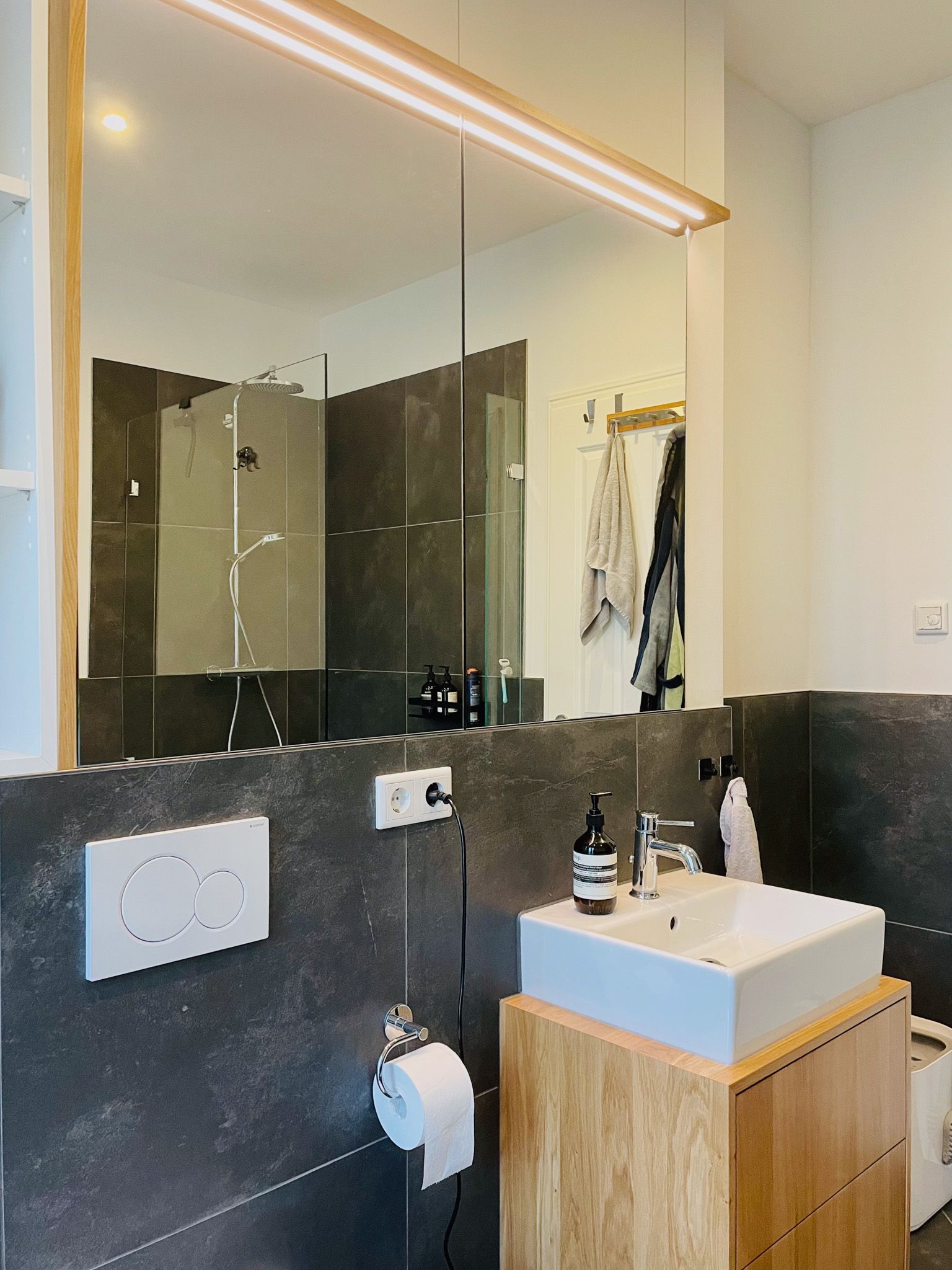 Modernes Badezimmer mit dunkelgrauen Fliesen, einem quadratischen Waschbecken auf einem Holzschrank und einem großen Spiegel.