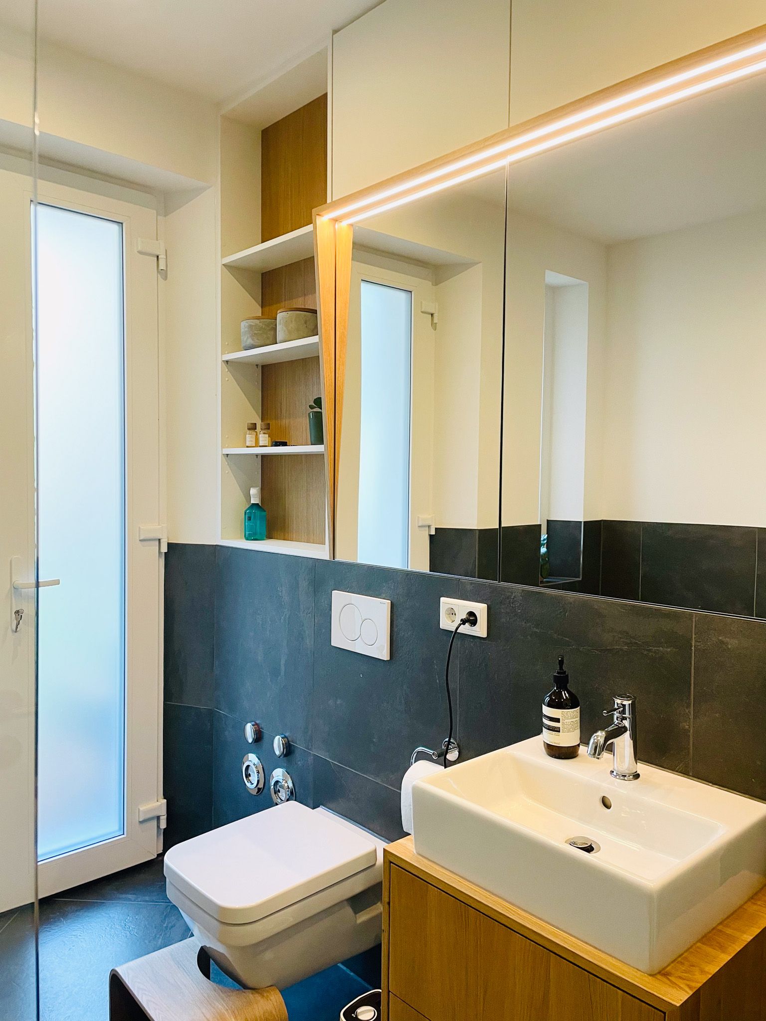 Modernes Badezimmer mit Toilette, Waschbecken, Spiegel und offenen Regalen. Farbschema in Grau und Weiß.