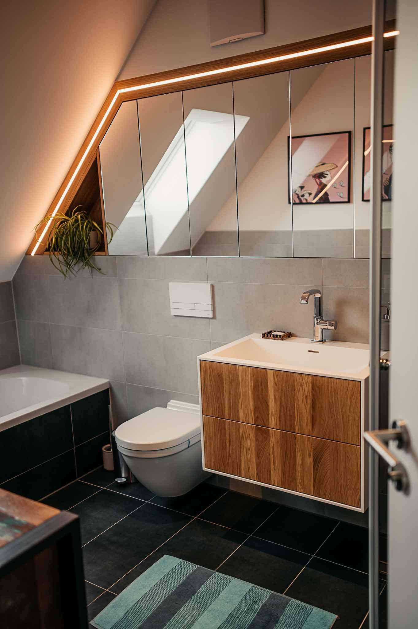 Modernes Badezimmer mit Holzwaschtisch, grauen Fliesen, schräger Decke und Toilette.