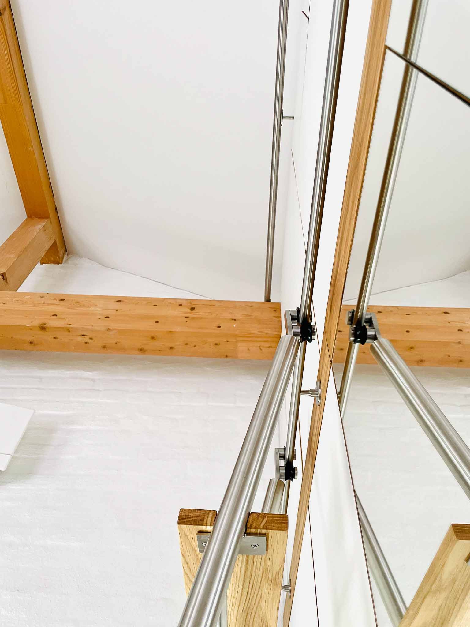 Klappbare Dachbodentreppe aus Holz mit Metallgeländer und Kette, an einer weißen Wand montiert, unter einer weißen Decke mit freiliegenden Holzbalken.