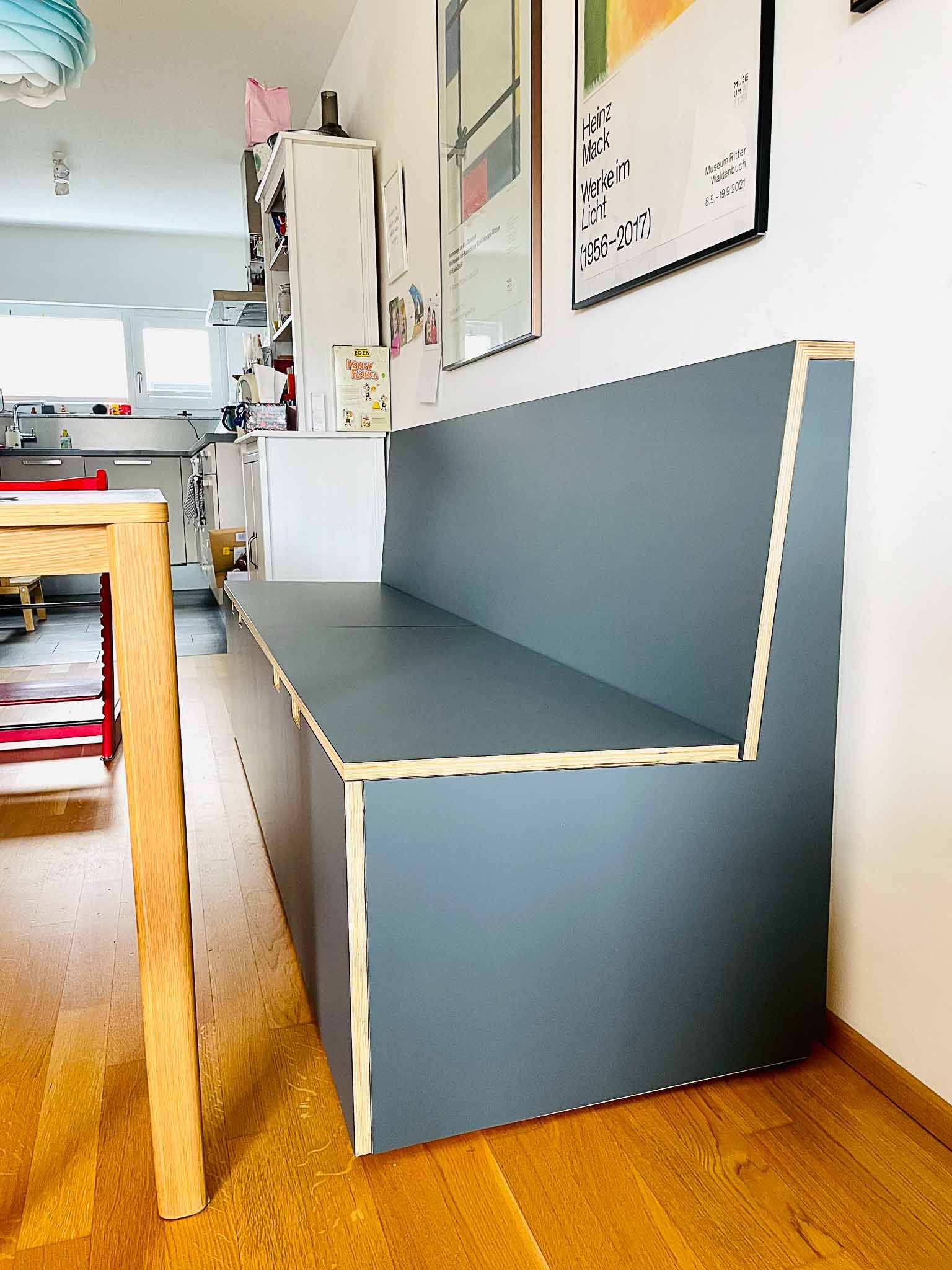 Graue Einbaubank mit Holzverkleidung vor einer weißen Wand, in einem Essbereich, neben einem Holztisch.
