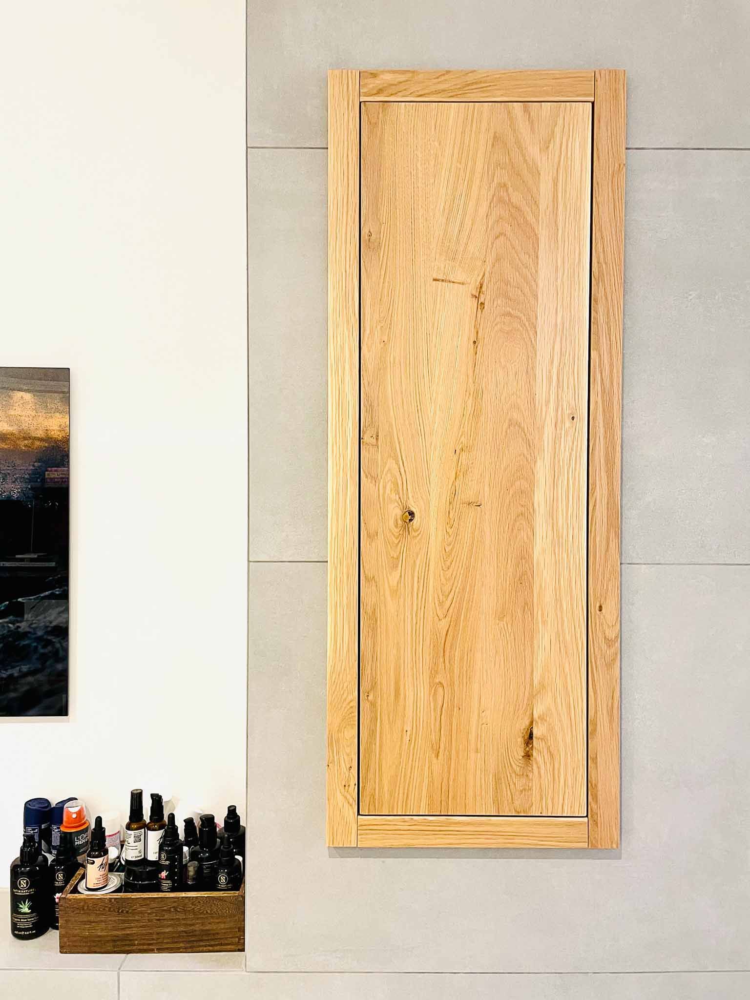 Holzschranktür, an einer grauen Wand montiert; auf der linken Seite befinden sich ein Gemälde und ein Regal mit Flaschen.