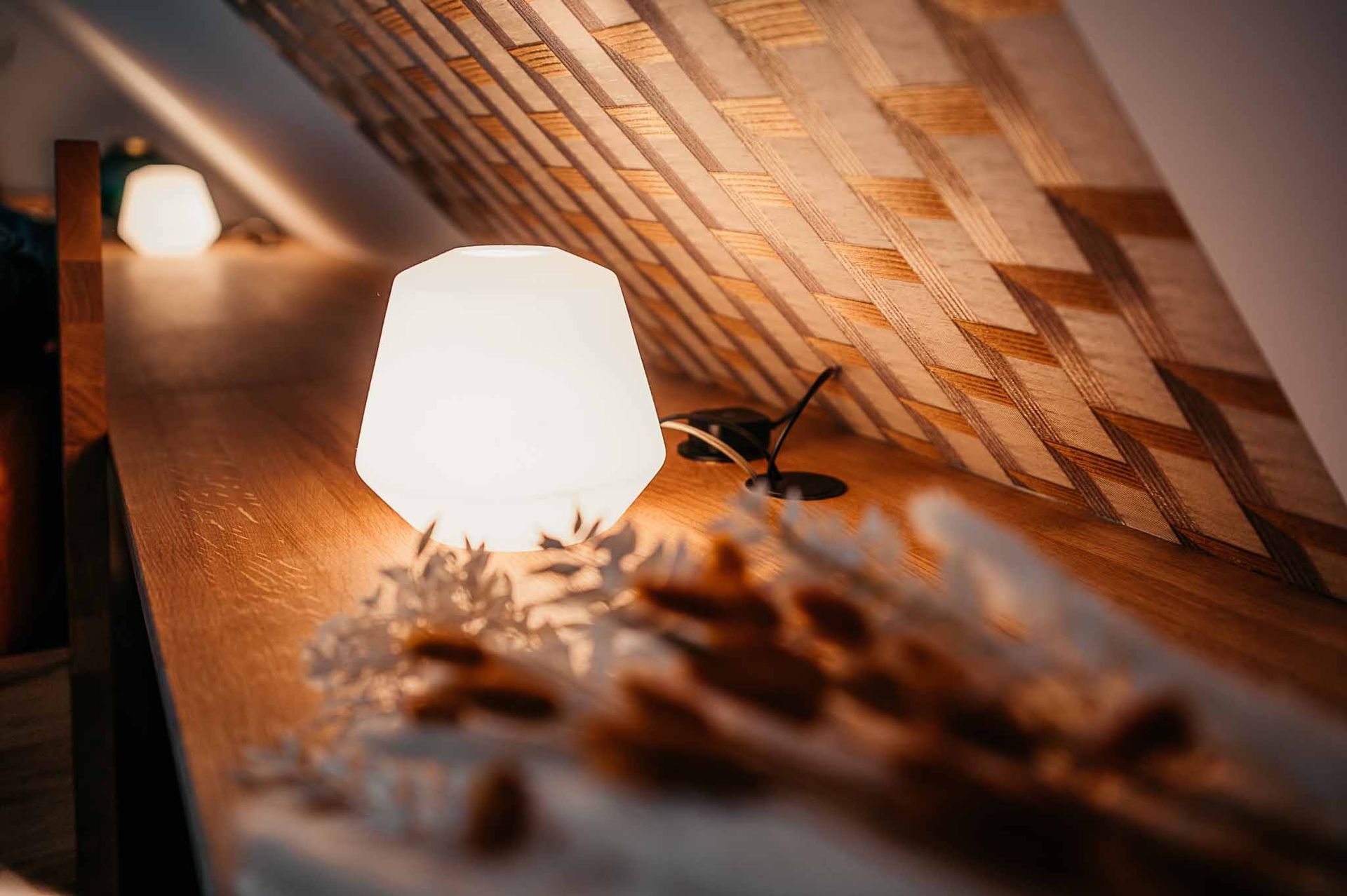 Eine warm leuchtende Lampe auf einem Holzregal beleuchtet getrocknete Blumen. Im Hintergrund eine diagonal strukturierte Wand.