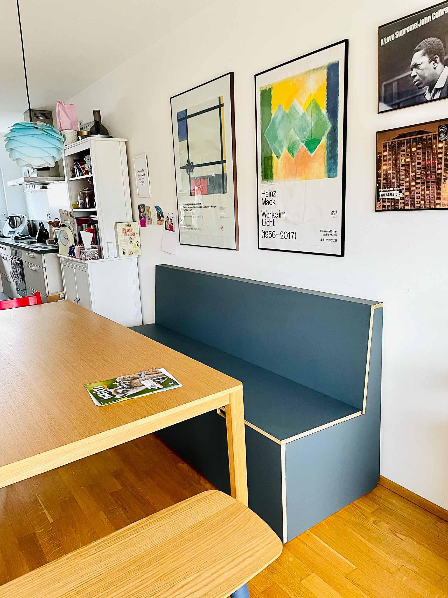 Ein Essbereich mit einem Holztisch und einer eingebauten blauen Bank. An der Wand hängt ein Kunstwerk.