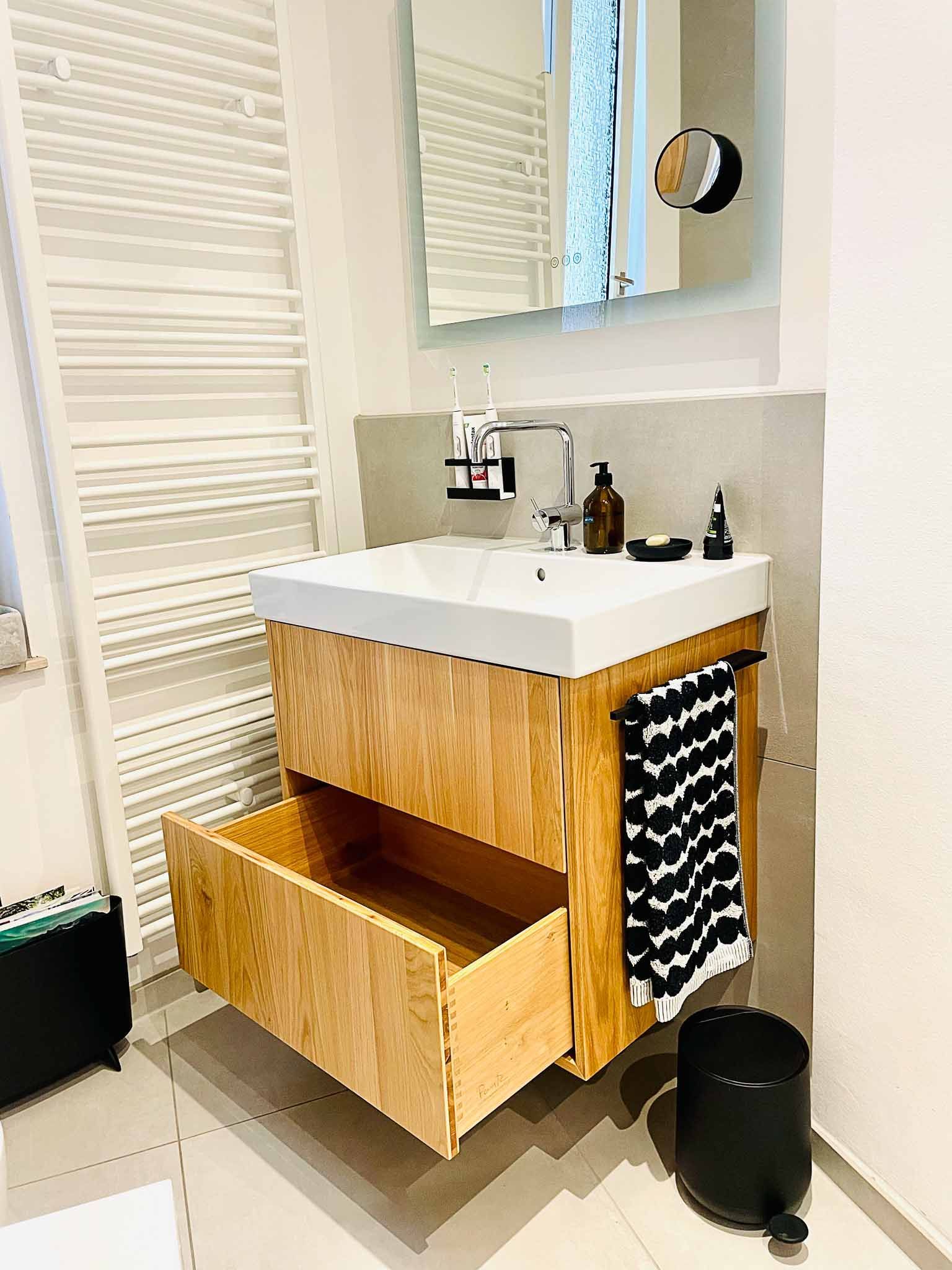 Badezimmer mit Waschtisch aus hellem Holz, offener Schublade, Waschbecken, Spiegel, Handtuch und Mülleimer.