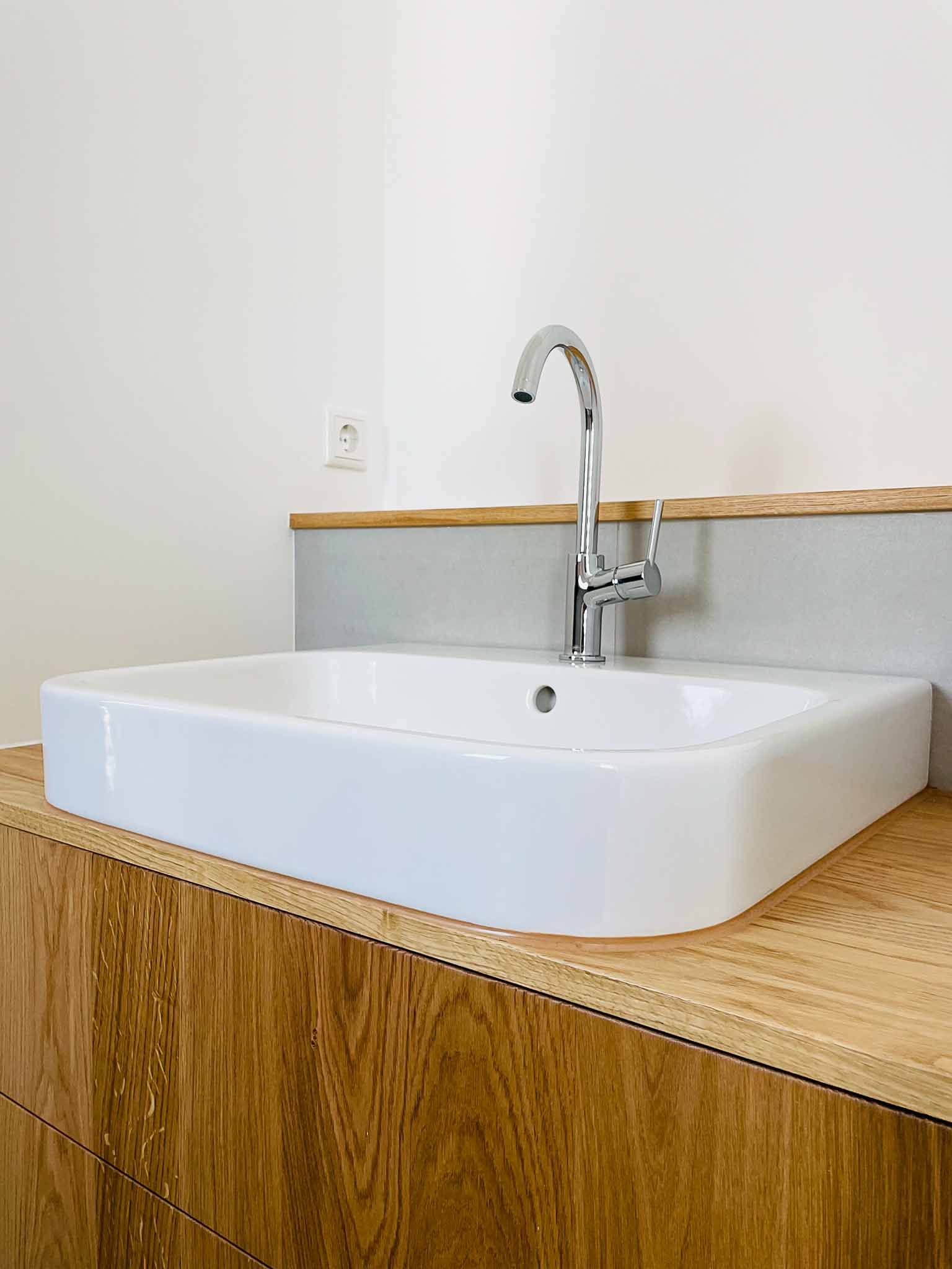 Weißes rechteckiges Waschbecken mit Chromhahn auf Holzschrank vor einer hellgrauen und weißen Wand.