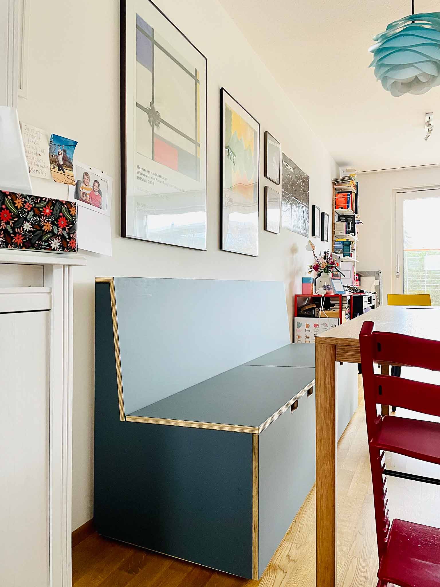 Eingebaute Bank mit blaugrauer Rückenlehne und Sitzfläche vor einer weißen Wand mit gerahmter Kunst. Essbereich.