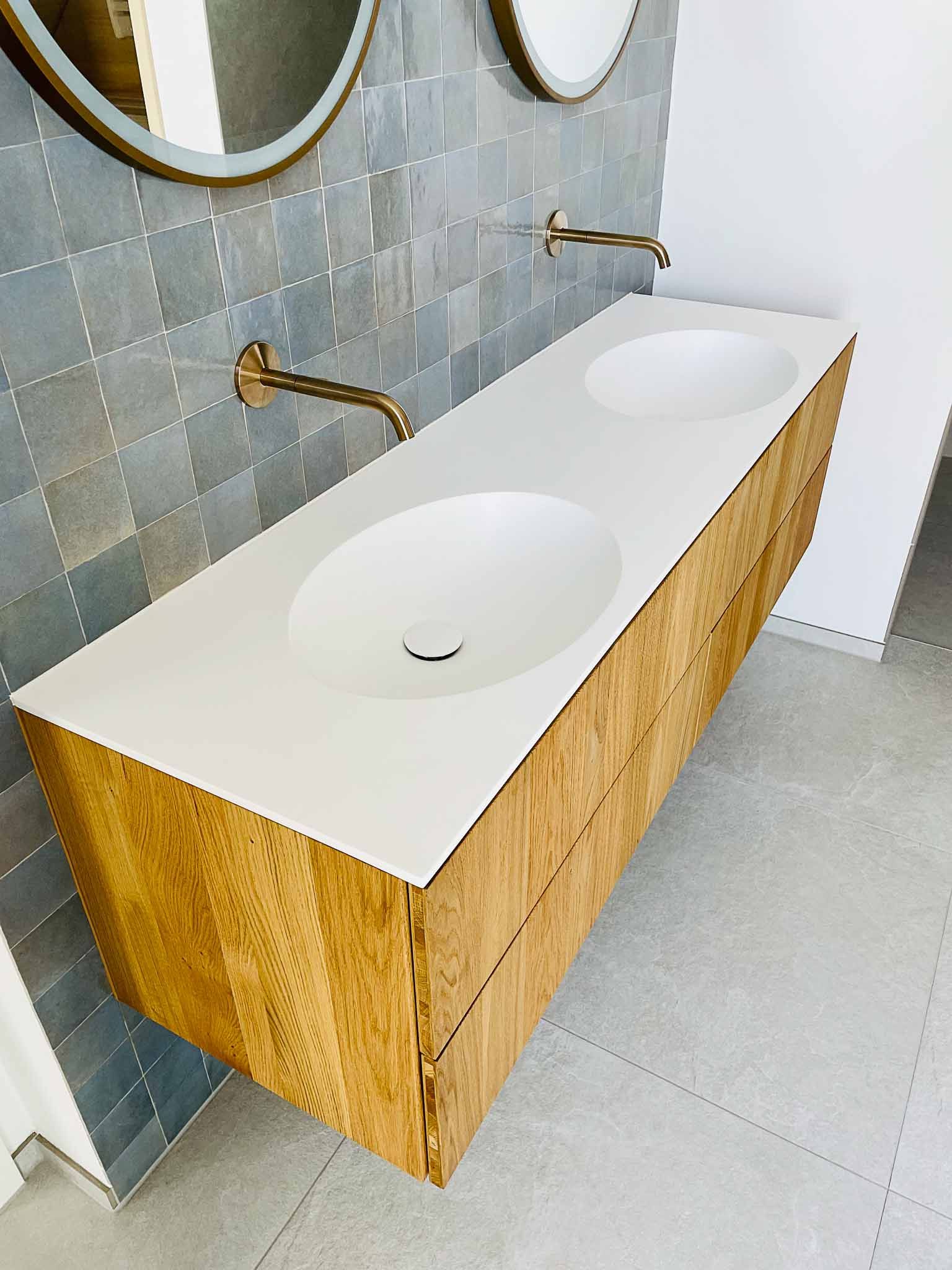 Schwebender Waschtisch aus Holz mit zwei weißen Waschbecken und goldenen Armaturen vor einer strukturierten grauen Fliesenwand.