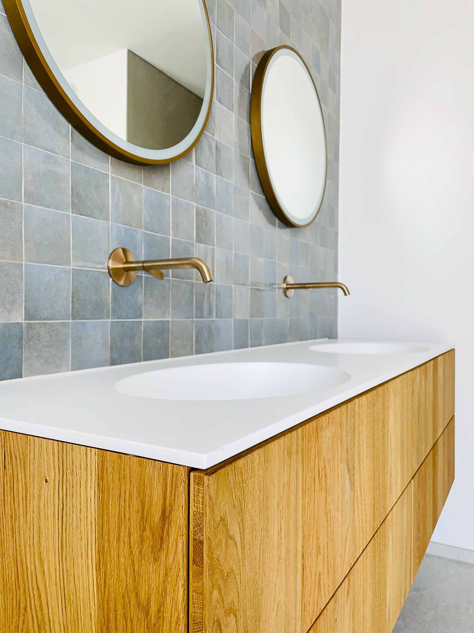 Holzwaschtisch mit weißen Waschbecken, goldenen Wasserhähnen und runden Spiegeln vor grauen Fliesen.