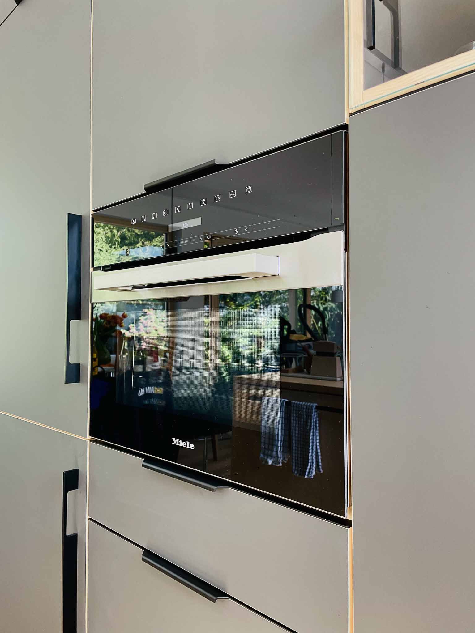 Schwarzer Ofen, eingebaut in hellgraue Schränke; auf dem Glas sind die Spiegelung einer Küche und von Grünpflanzen im Freien sichtbar.