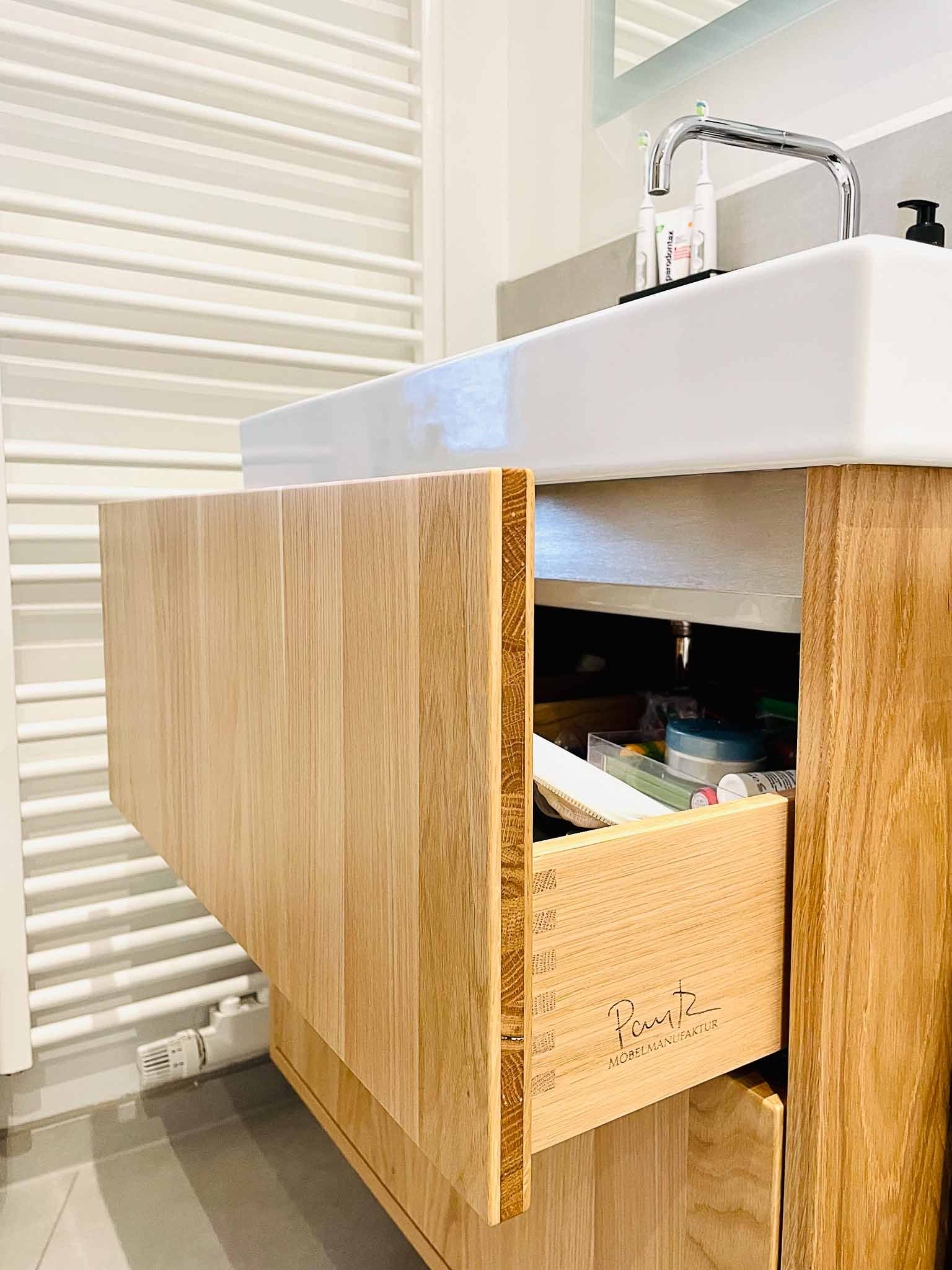 Badezimmerwaschtisch mit offener Holzschublade, in der Toilettenartikel zum Vorschein kommen. Heller Holzschrank mit weißem Waschbecken.