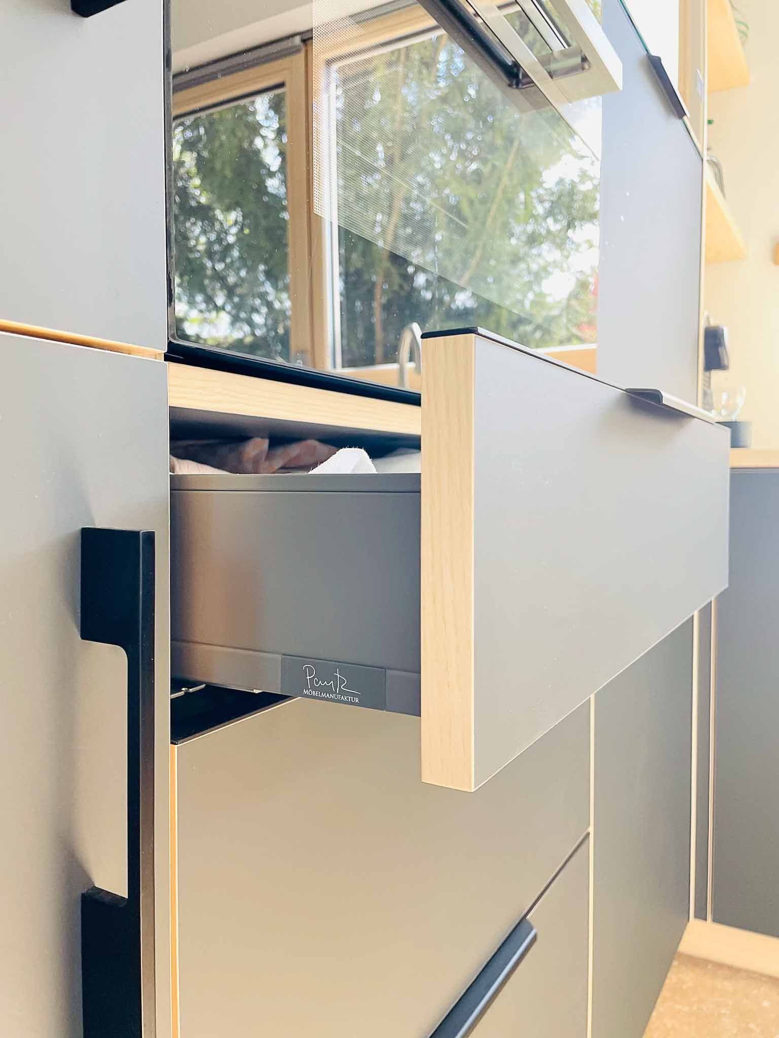 Offene graue Küchenschublade mit dunklem Griff, heller Holzverkleidung, Fenster im Hintergrund.