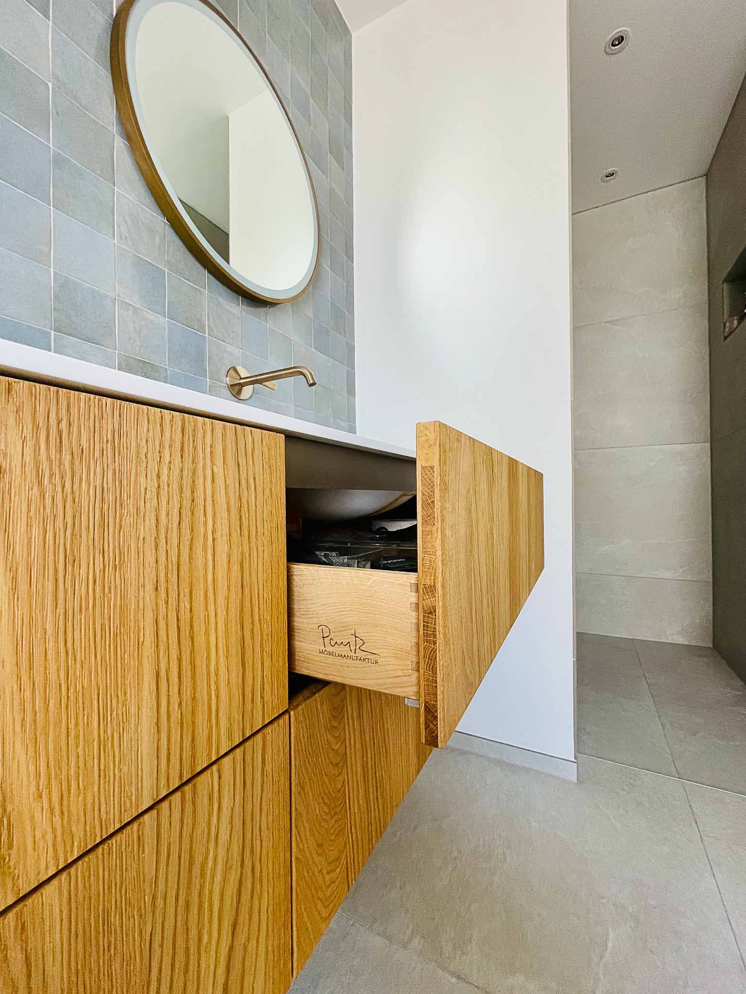 Badezimmerschrank aus Holz mit offener Schublade, goldenem Spiegel und blau gefliester Wand.