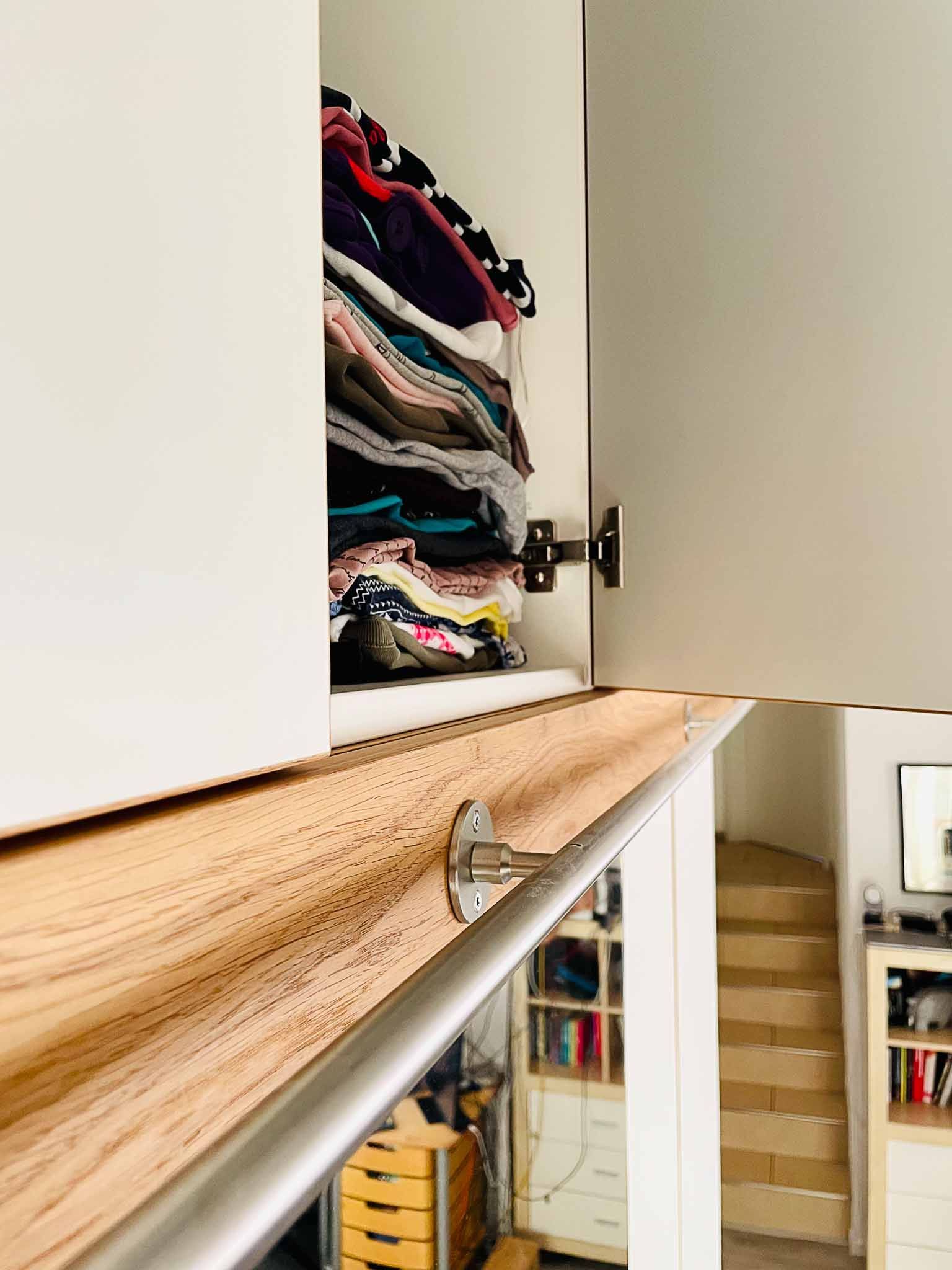 Schranktür geöffnet, darunter ein Kleiderstapel. Geländer aus Holz und Metall; Stufen im Hintergrund.