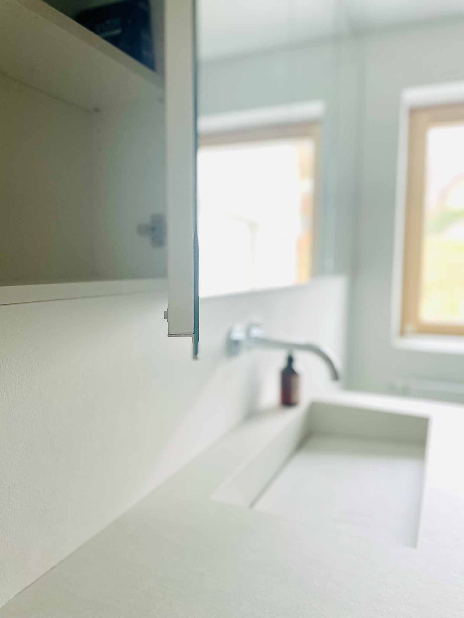 Badezimmer mit weißem Waschbecken, Wasserhahn, Spiegel und Schrank. Sonnenlicht fällt durch die Fenster.