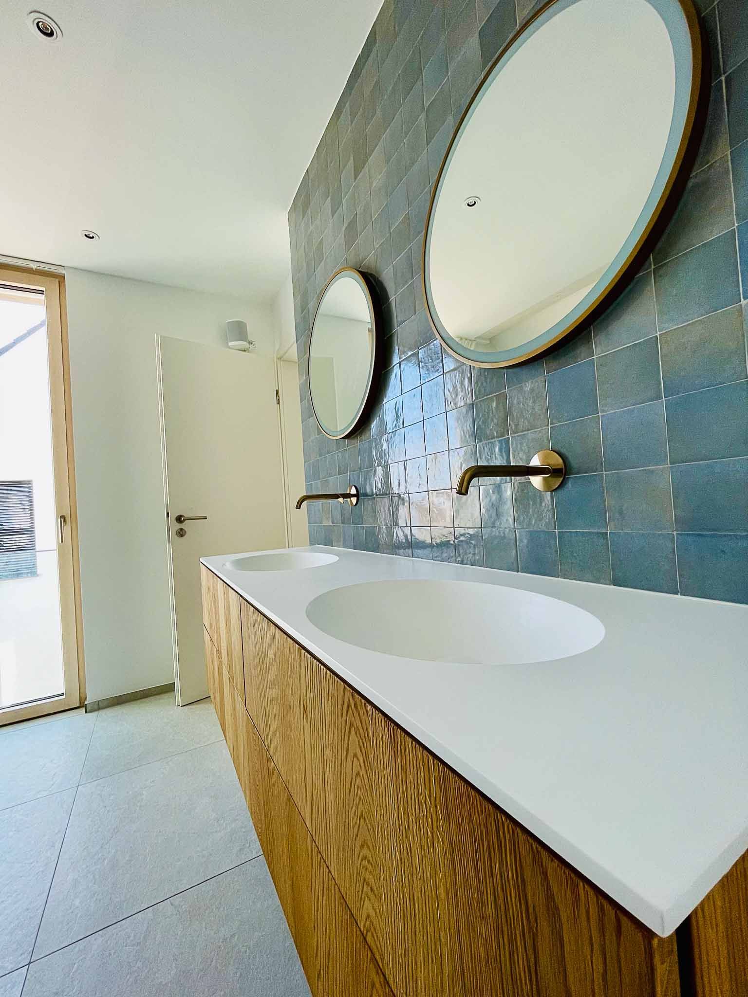 Modernes Badezimmer mit Holzwaschtisch, weißer Arbeitsplatte mit zwei Waschbecken, blau gefliester Wand und zwei runden Spiegeln.