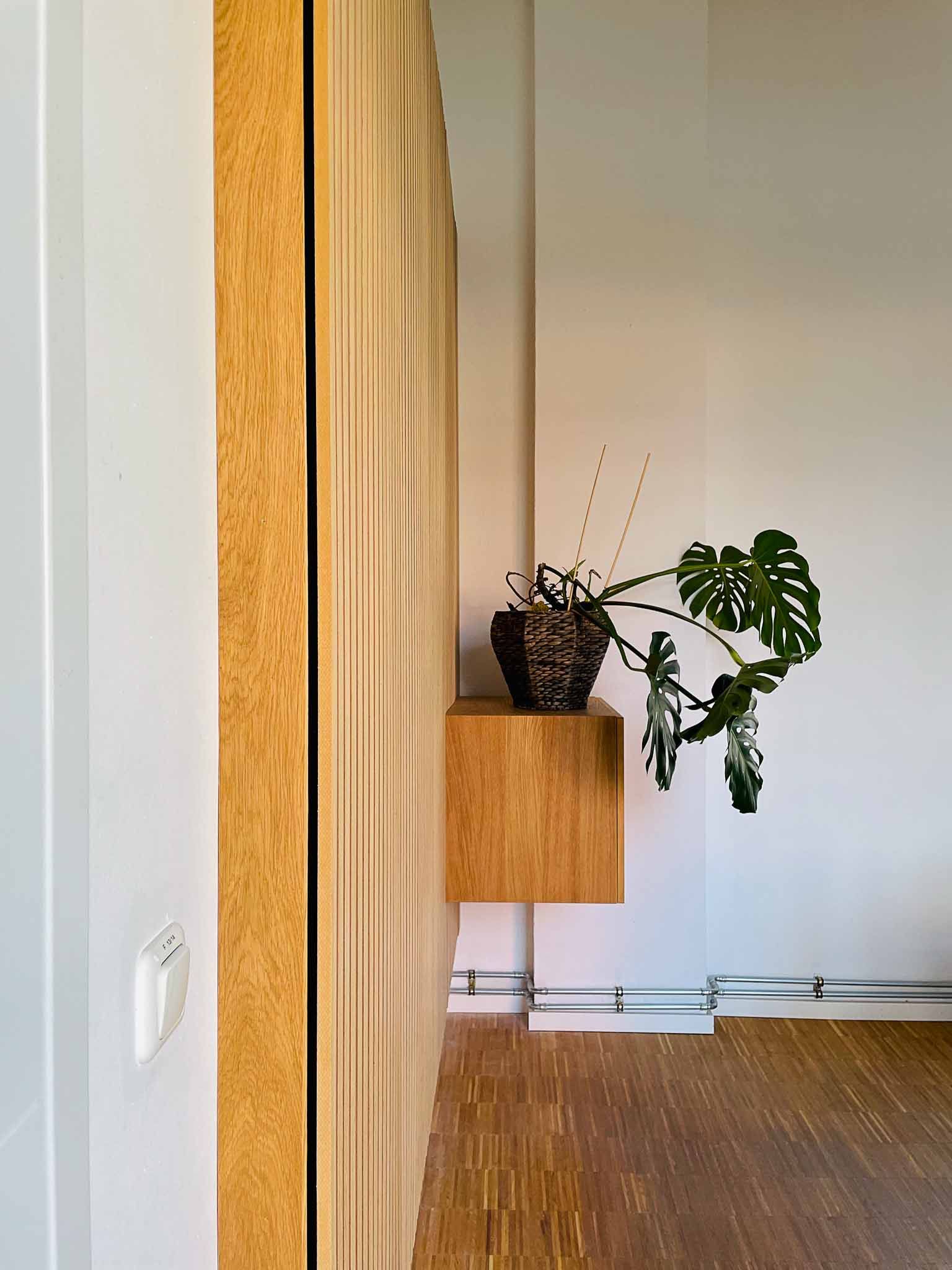 Strukturierte Holzwand mit einem schwebenden Holzregal, auf dem eine Topfpflanze steht. Brauner Boden, weiße Wände.