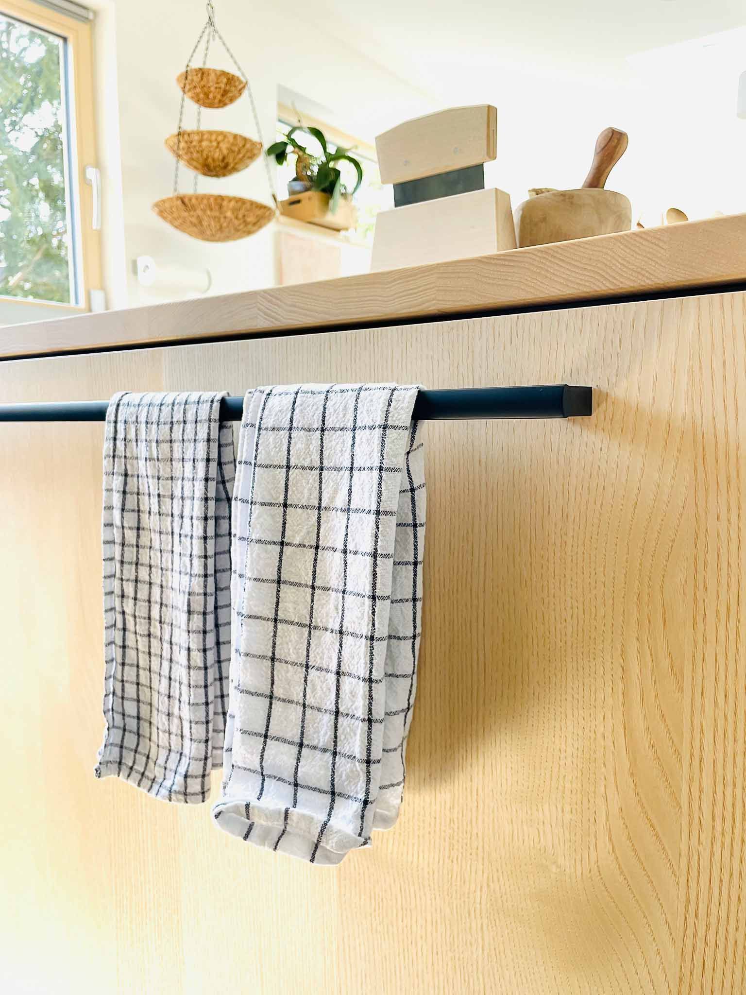 Zwei weiß karierte Küchentücher hängen an einer schwarzen Stange auf einem hellen Holzschrank.