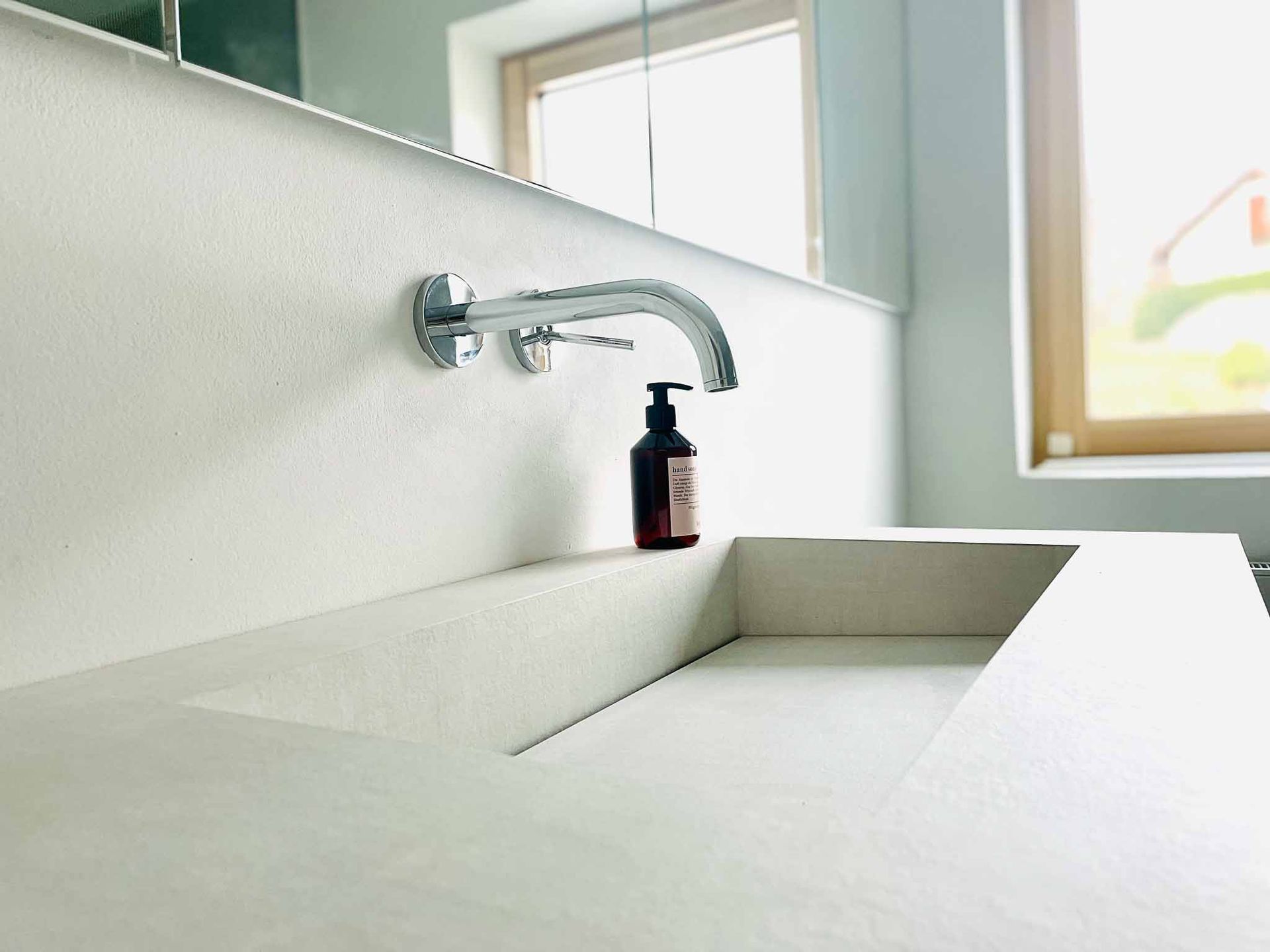 Weißes Waschbecken mit Wandarmatur. Braune Seifenflasche auf dem Sims, Fenster im Hintergrund.